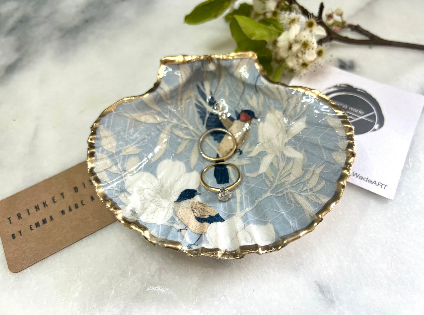 Handmade scallop shell trinket dish featuring a Blue Birds of Paradise decoupage design, finished with real gold leaf and sealed with glossy resin. Crafted from a natural Dorset scallop shell and shown as a jewellery and ring holder on a light background. Coastal-inspired home decor, perfect for holding rings, earrings and small keepsakes, with each shell one of a kind and finished by hand.