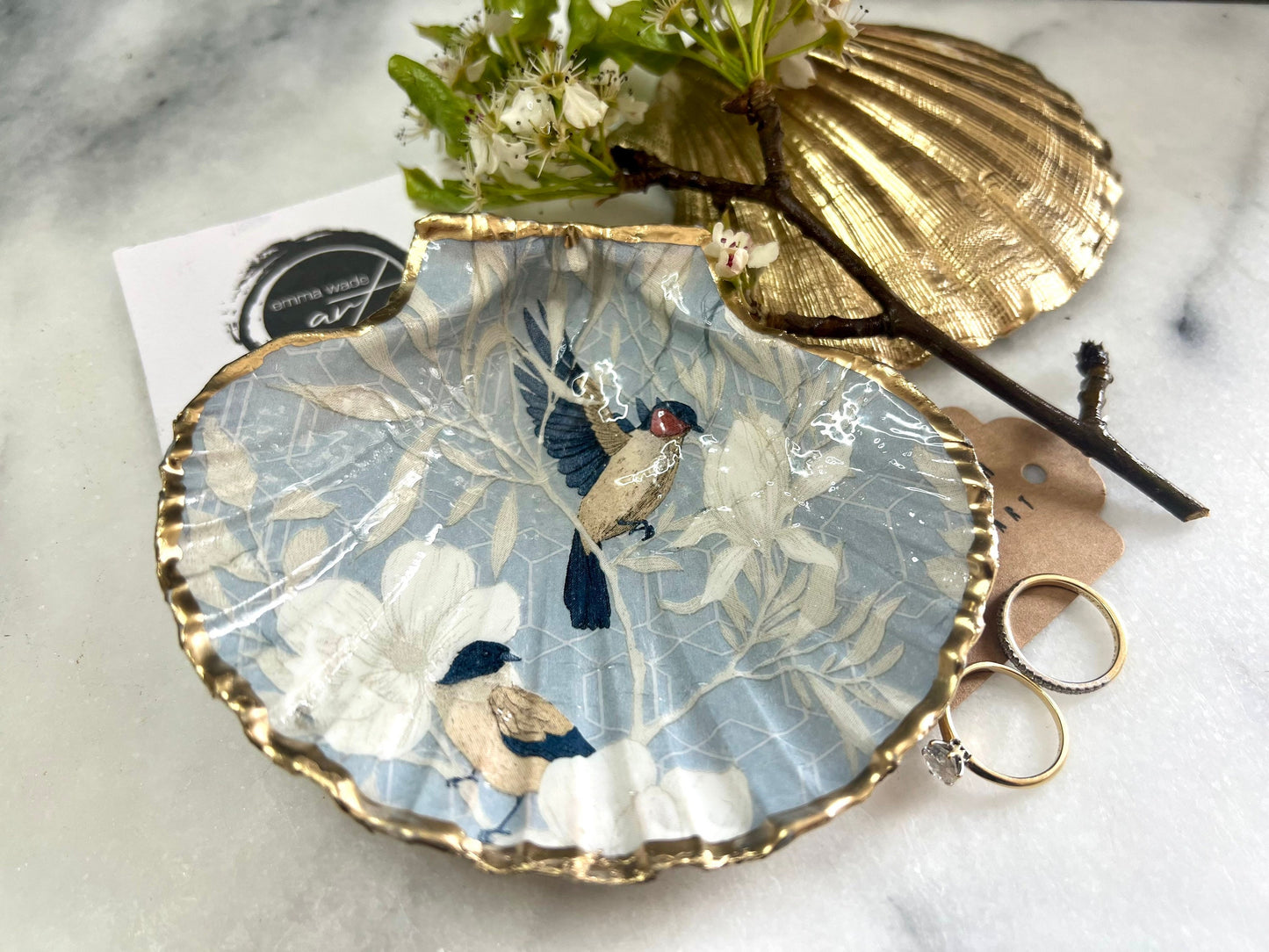 Handmade scallop shell trinket dish featuring a Blue Birds of Paradise decoupage design, finished with real gold leaf and sealed with glossy resin. Crafted from a natural Dorset scallop shell and shown as a jewellery and ring holder on a light background. Coastal-inspired home decor, perfect for holding rings, earrings and small keepsakes, with each shell one of a kind and finished by hand.