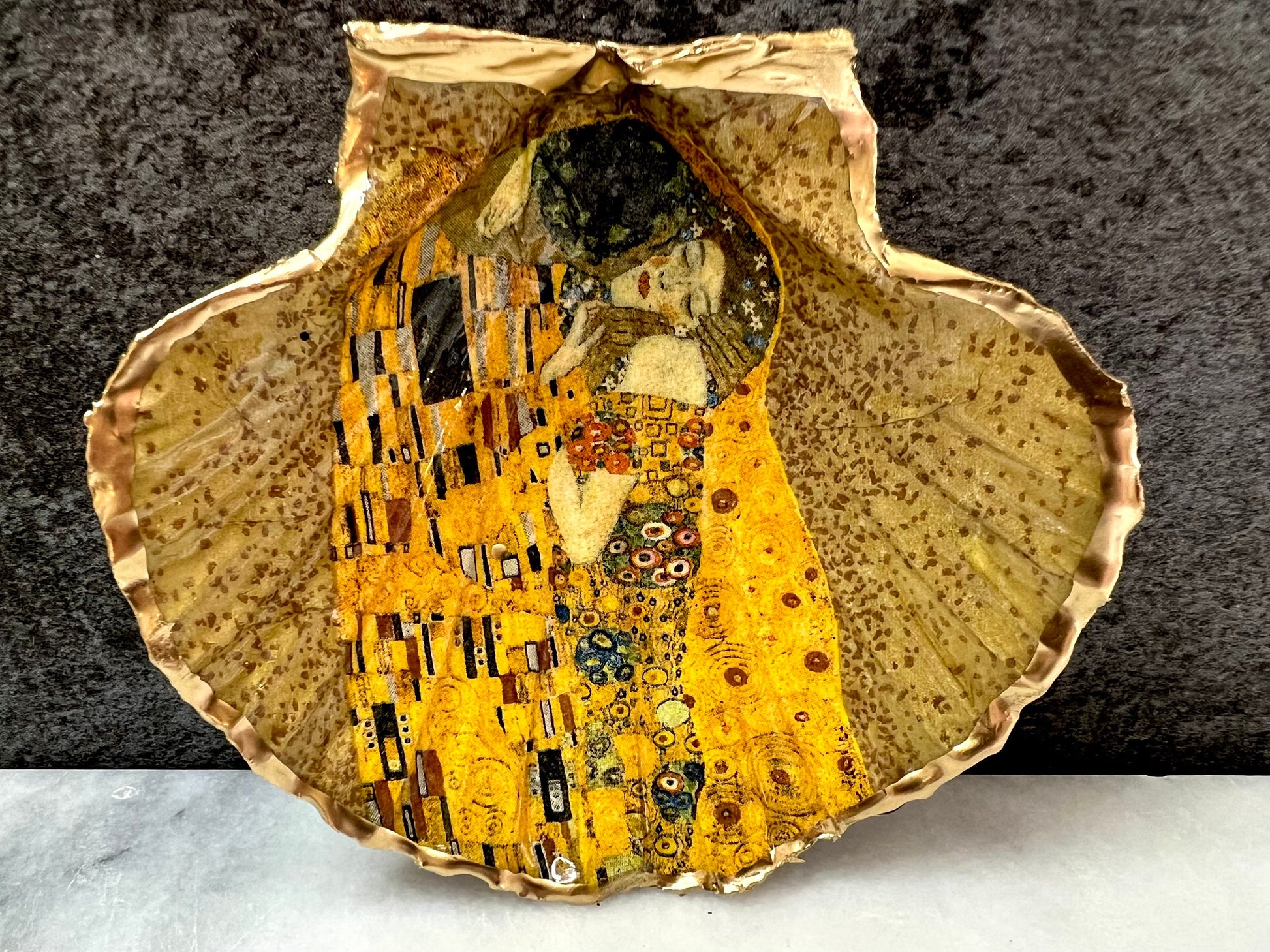 Handmade scallop shell trinket dish inspired by Gustav Klimt’s “The Kiss”, finished with gold leaf and sealed with glossy resin. Coastal Dorset shell jewellery dish perfect for rings, earrings and small treasures. Unique decorative shell bowl with golden artwork design, photographed on a light background showing detailed gilded surface and natural shell shape.