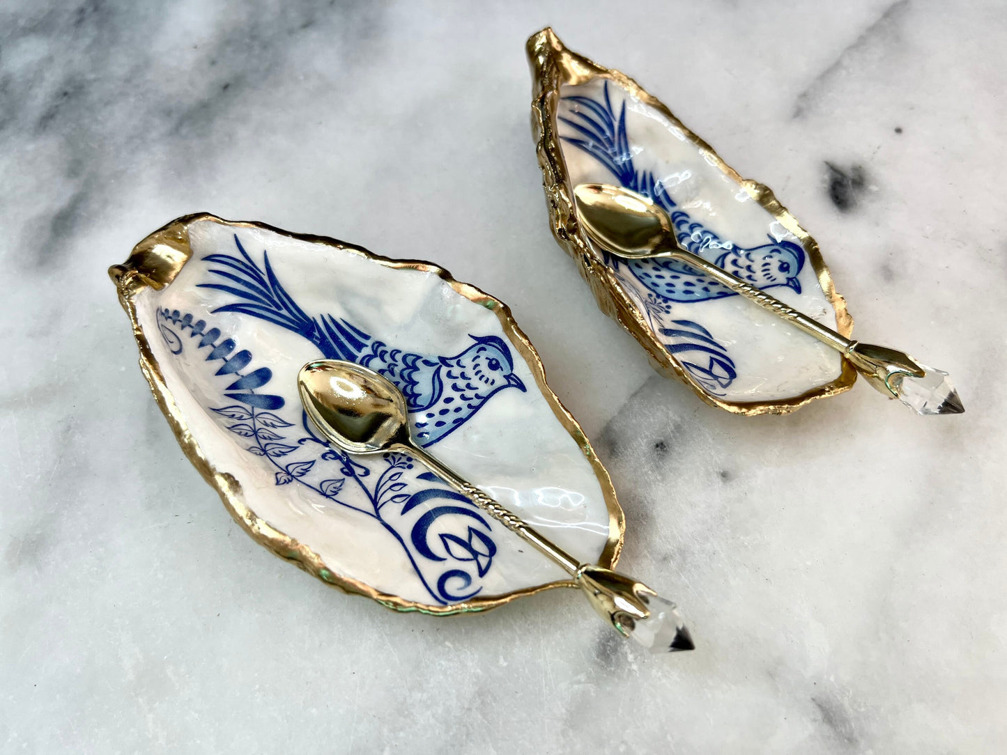 Handmade blue bird oyster shell salt and pepper pinch pot set crafted from natural Dorset oyster shells with glossy pearlescent interiors, blue porcelain style bird artwork and shimmering liquid gold leaf edging. Luxury coastal cruet dishes sealed with food-safe resin and styled with optional gold spoons. Elegant seaside dining decor for beach house kitchens, dinner parties, weddings, anniversaries and bird lovers. Artisan shell tableware handmade in the UK with coastal blue and white styling.