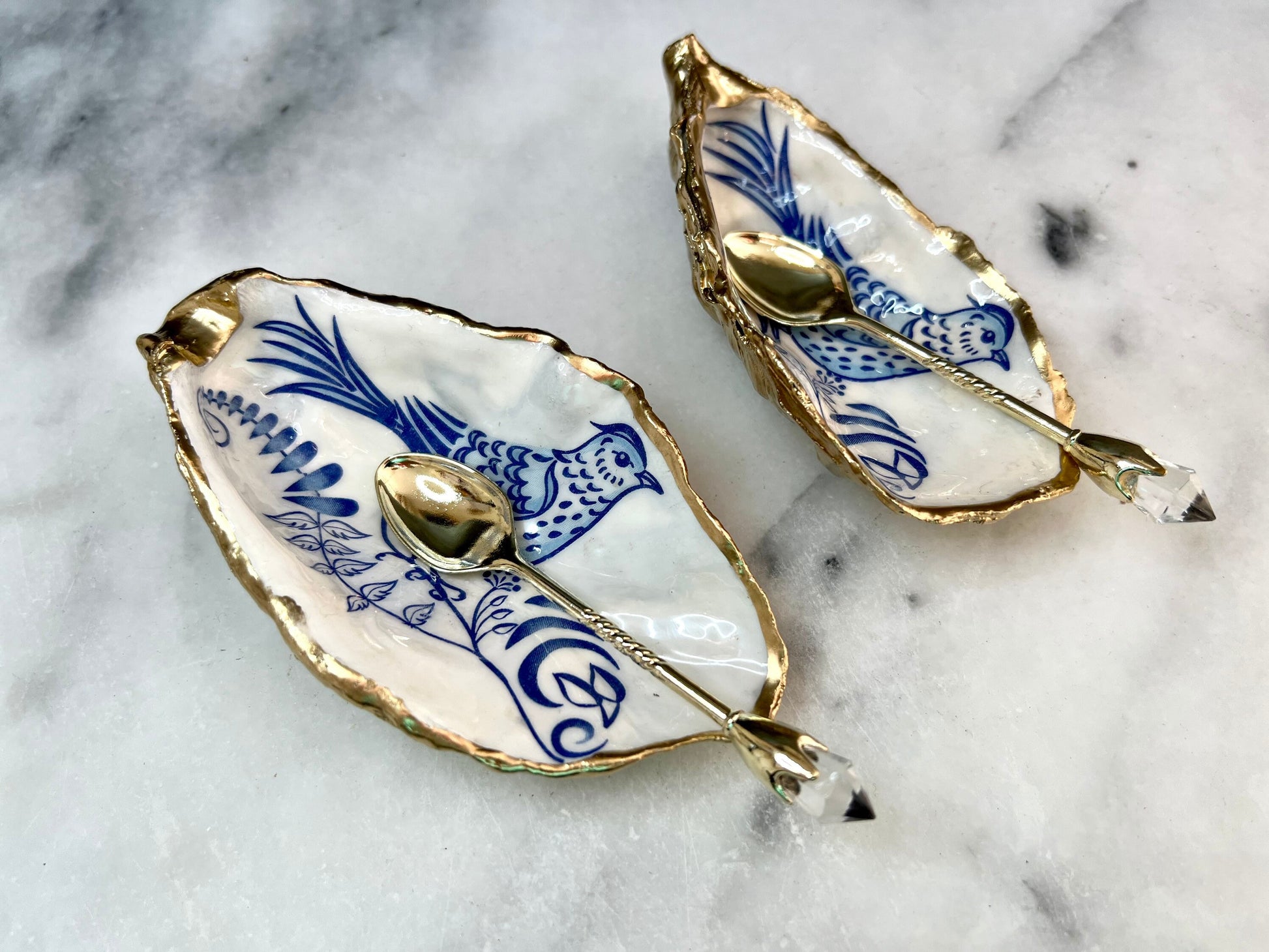 Handmade blue bird oyster shell salt and pepper pinch pot set crafted from natural Dorset oyster shells with glossy pearlescent interiors, blue porcelain style bird artwork and shimmering liquid gold leaf edging. Luxury coastal cruet dishes sealed with food-safe resin and styled with optional gold spoons. Elegant seaside dining decor for beach house kitchens, dinner parties, weddings, anniversaries and bird lovers. Artisan shell tableware handmade in the UK with coastal blue and white styling.