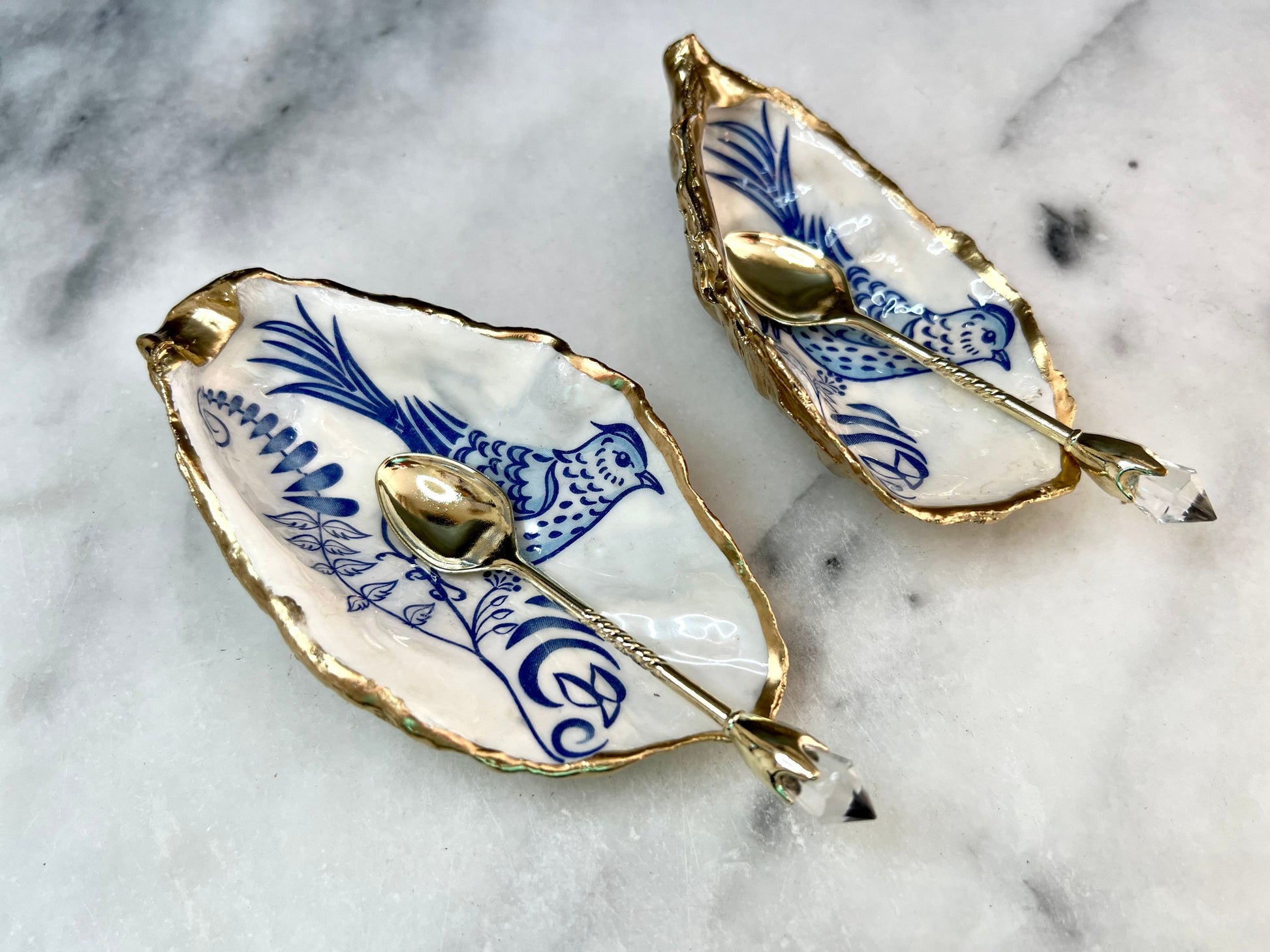Handmade blue bird oyster shell salt and pepper pinch pot set crafted from natural Dorset oyster shells with glossy pearlescent interiors, blue porcelain style bird artwork and shimmering liquid gold leaf edging. Luxury coastal cruet dishes sealed with food-safe resin and styled with optional gold spoons. Elegant seaside dining decor for beach house kitchens, dinner parties, weddings, anniversaries and bird lovers. Artisan shell tableware handmade in the UK with coastal blue and white styling.