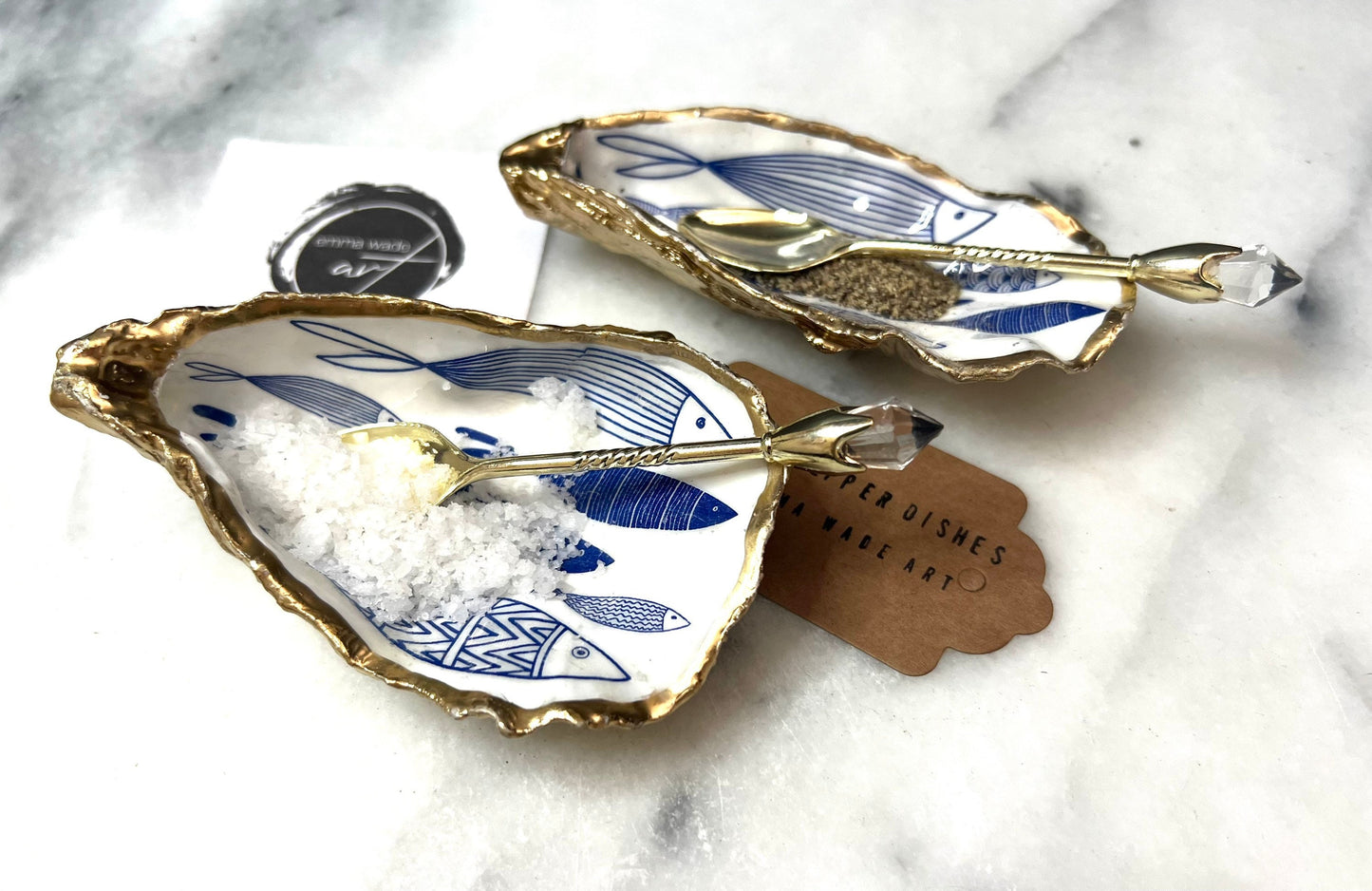Pair of handmade Dorset oyster shell salt and pepper pinch pots decorated with elegant blue and white fish artwork, finished with hand applied liquid gold leaf edging and sealed in glossy food-safe resin. Coastal shell cruet dishes styled with optional gold crystal spoons on a marble background. Luxury nautical kitchen decor inspired by the sea, perfect for beach house dining, dinner parties, weddings, anniversaries, and coastal home gifts.