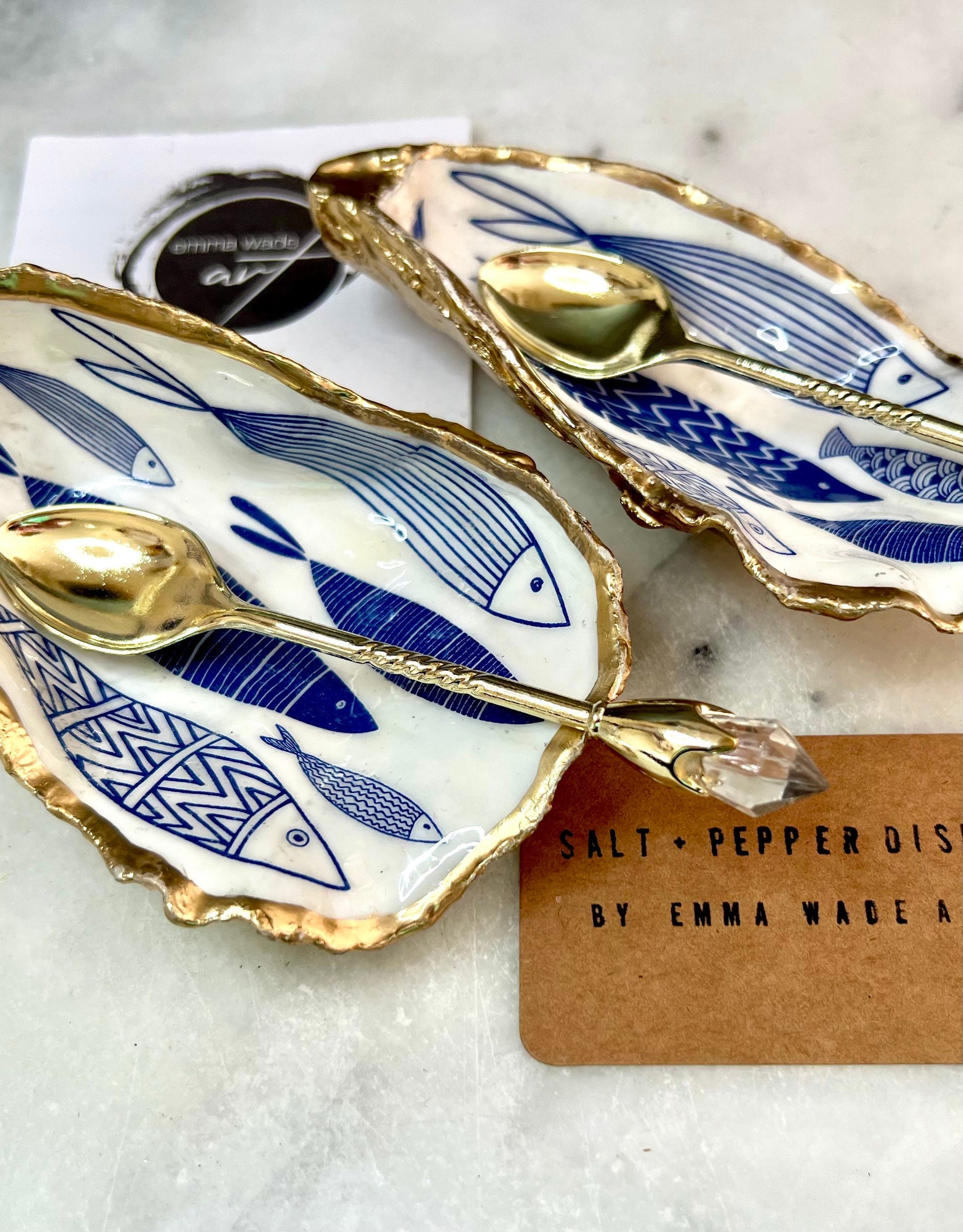 Pair of handmade Dorset oyster shell salt and pepper pinch pots decorated with elegant blue and white fish artwork, finished with hand applied liquid gold leaf edging and sealed in glossy food-safe resin. Coastal shell cruet dishes styled with optional gold crystal spoons on a marble background. Luxury nautical kitchen decor inspired by the sea, perfect for beach house dining, dinner parties, weddings, anniversaries, and coastal home gifts.