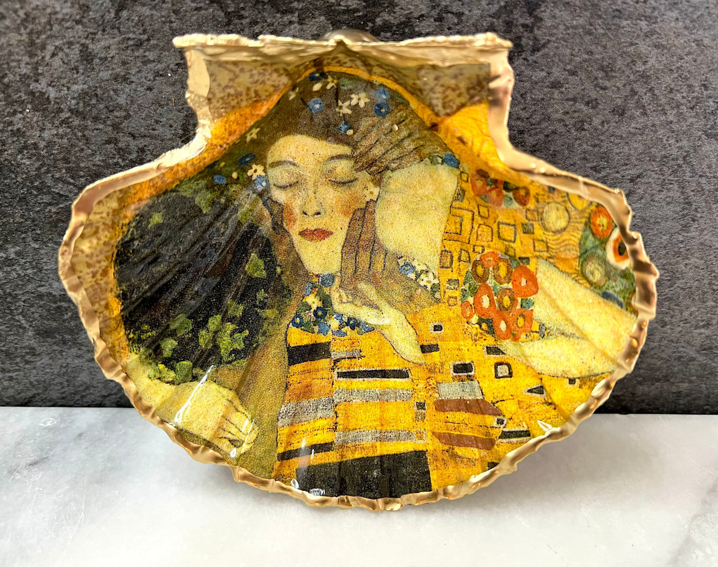 Handmade scallop shell trinket dish inspired by Gustav Klimt’s “The Kiss”, finished with gold leaf and sealed with glossy resin. Coastal Dorset shell jewellery dish perfect for rings, earrings and small treasures. Unique decorative shell bowl with golden artwork design, photographed on a light background showing detailed gilded surface and natural shell shape.
