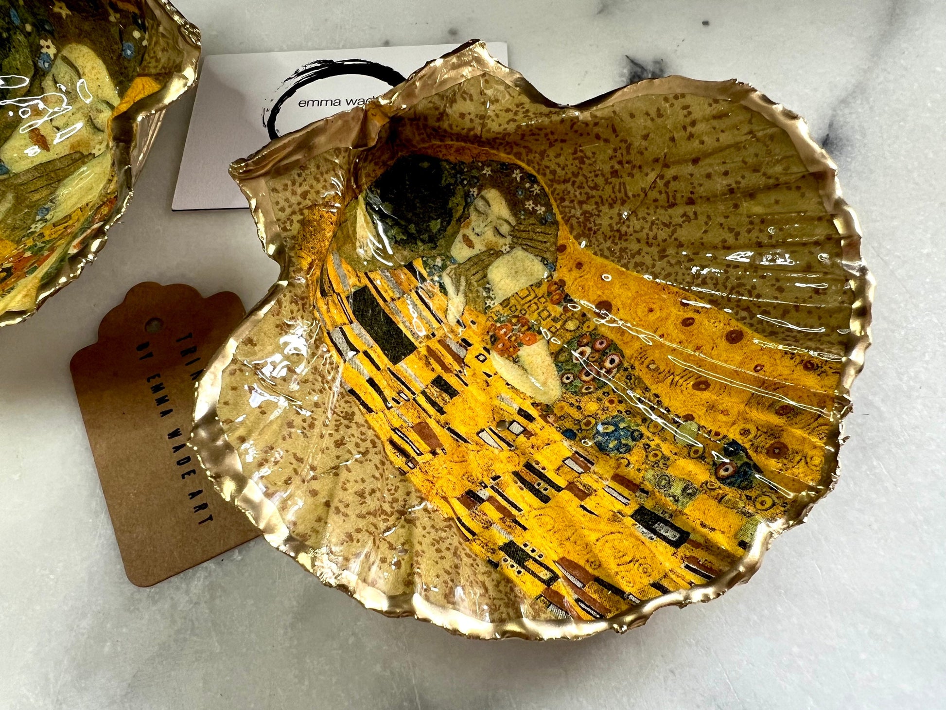 Handmade scallop shell trinket dish inspired by Gustav Klimt’s “The Kiss”, finished with gold leaf and sealed with glossy resin. Coastal Dorset shell jewellery dish perfect for rings, earrings and small treasures. Unique decorative shell bowl with golden artwork design, photographed on a light background showing detailed gilded surface and natural shell shape.