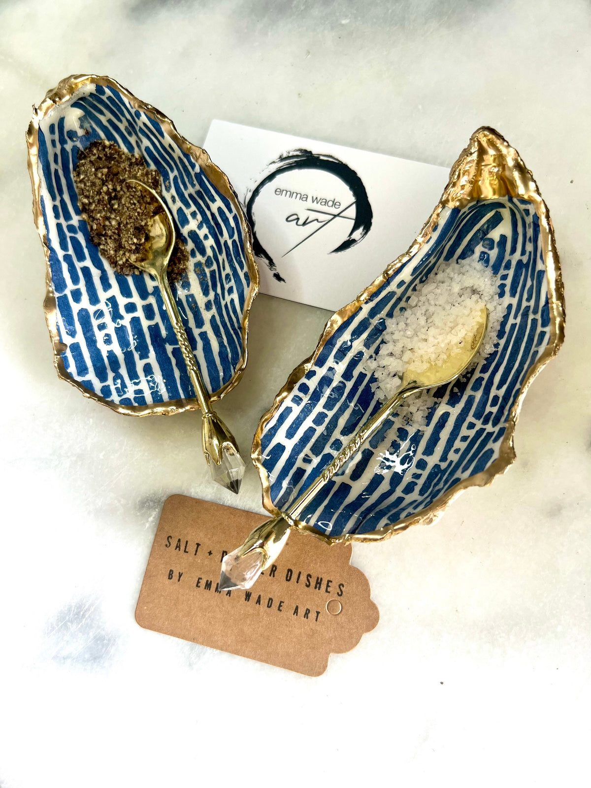 Pair of handmade blue and white lined oyster shell salt and pepper pinch pots with gold leaf trim, shown on a light background with small gold spoons. Crafted from natural Dorset oyster shells and sealed with glossy, food-safe resin, these coastal-inspired shell dishes are designed for table styling, weddings, or a unique hostess gift. Each shell is one of a kind and finished by hand.