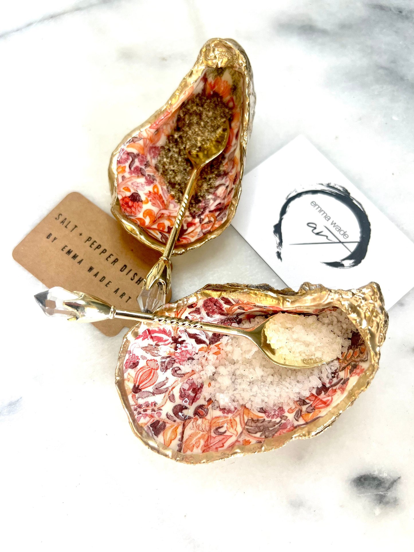 Handmade Dorset oyster shell salt and pepper pinch pots featuring a pink William Morris inspired floral design with hand-painted gold leaf edging. Decorative coastal tableware sealed in glossy food-safe resin and styled with gold crystal spoons on a marble background.