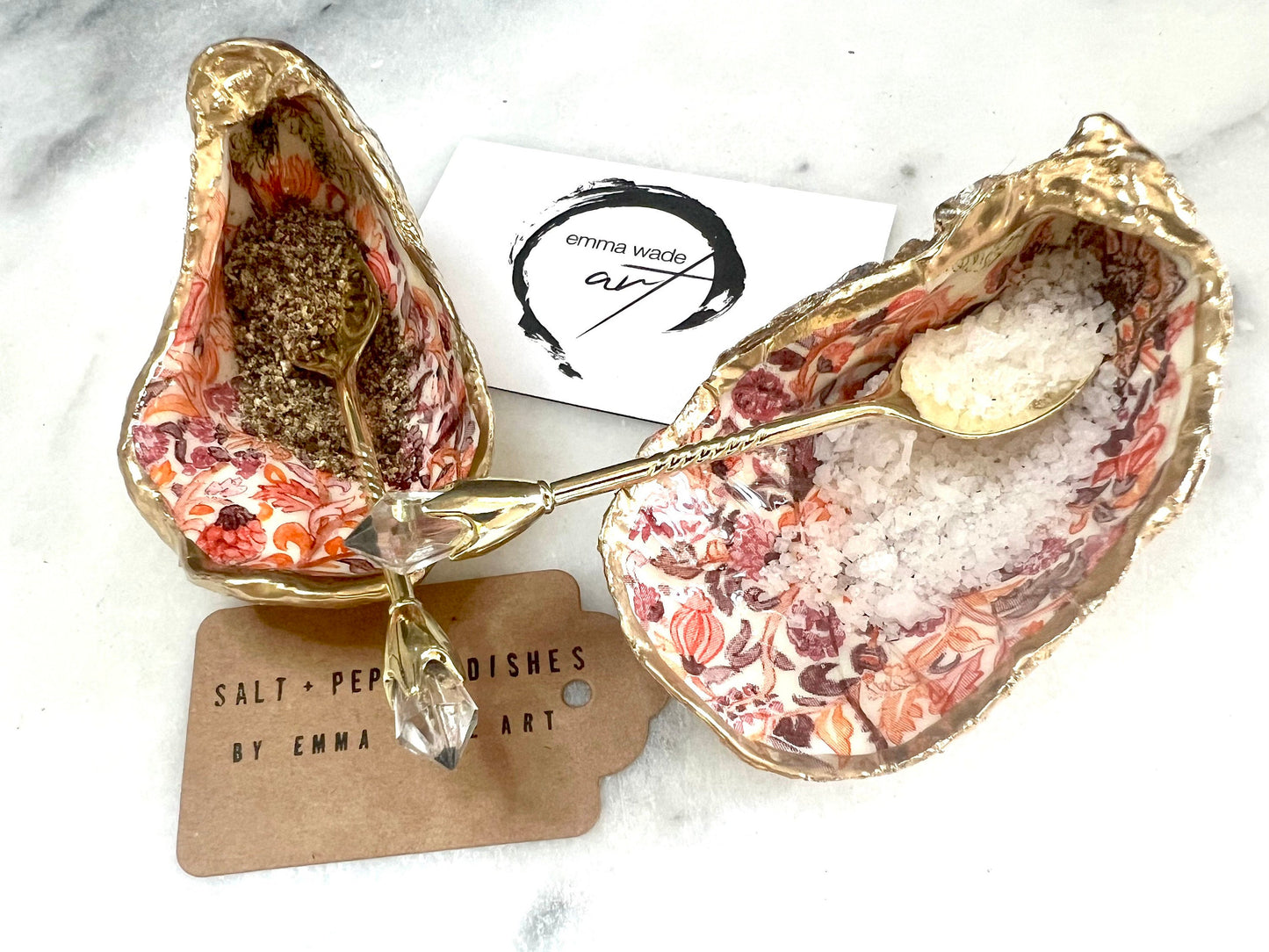 Handmade Dorset oyster shell salt and pepper pinch pots featuring a pink William Morris inspired floral design with hand-painted gold leaf edging. Decorative coastal tableware sealed in glossy food-safe resin and styled with gold crystal spoons on a marble background.