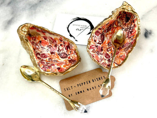 Handmade Dorset oyster shell salt and pepper pinch pots featuring a pink William Morris inspired floral design with hand-painted gold leaf edging. Decorative coastal tableware sealed in glossy food-safe resin and styled with gold crystal spoons on a marble background.