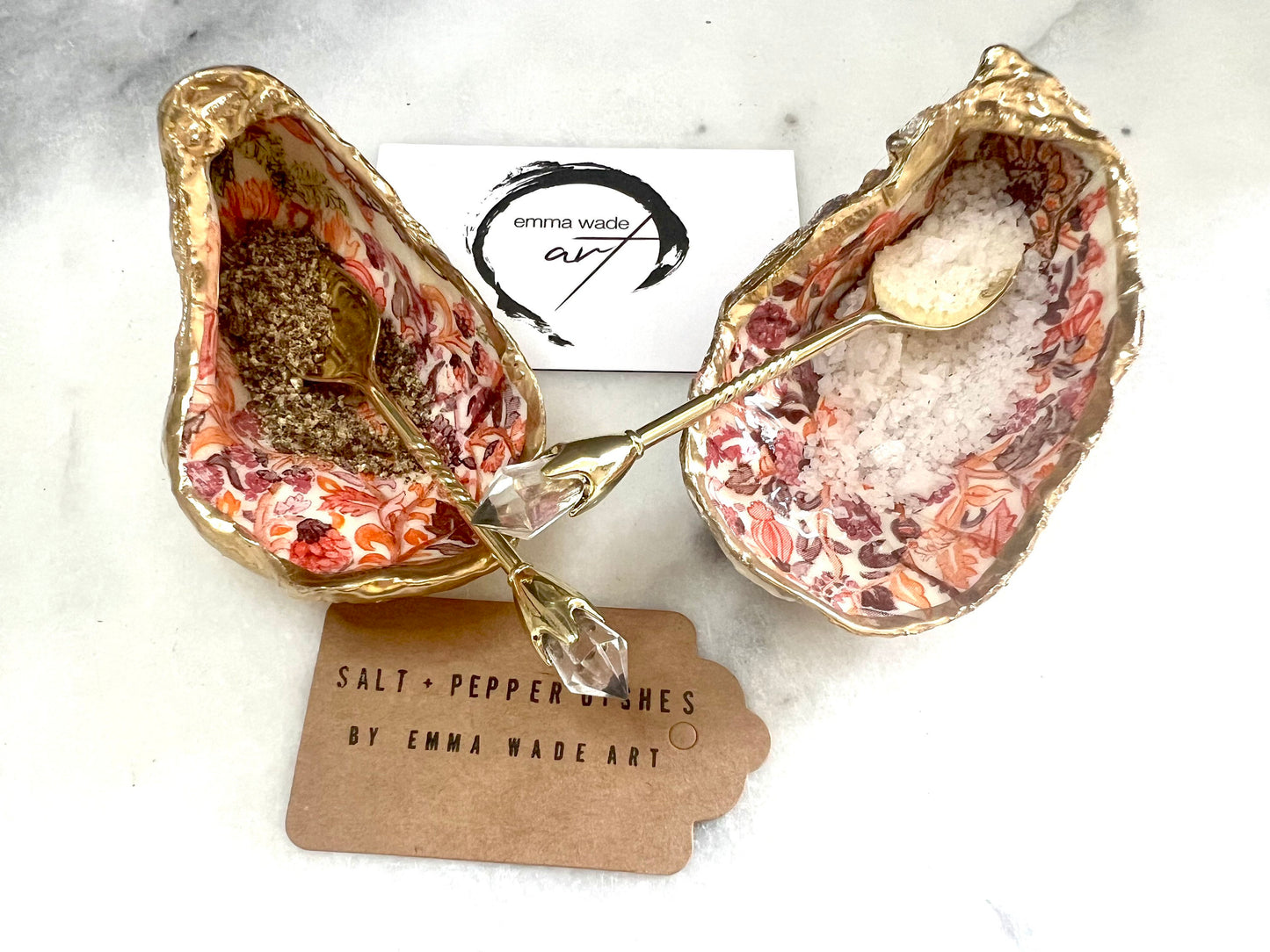 Handmade Dorset oyster shell salt and pepper pinch pots featuring a pink William Morris inspired floral design with hand-painted gold leaf edging. Decorative coastal tableware sealed in glossy food-safe resin and styled with gold crystal spoons on a marble background.