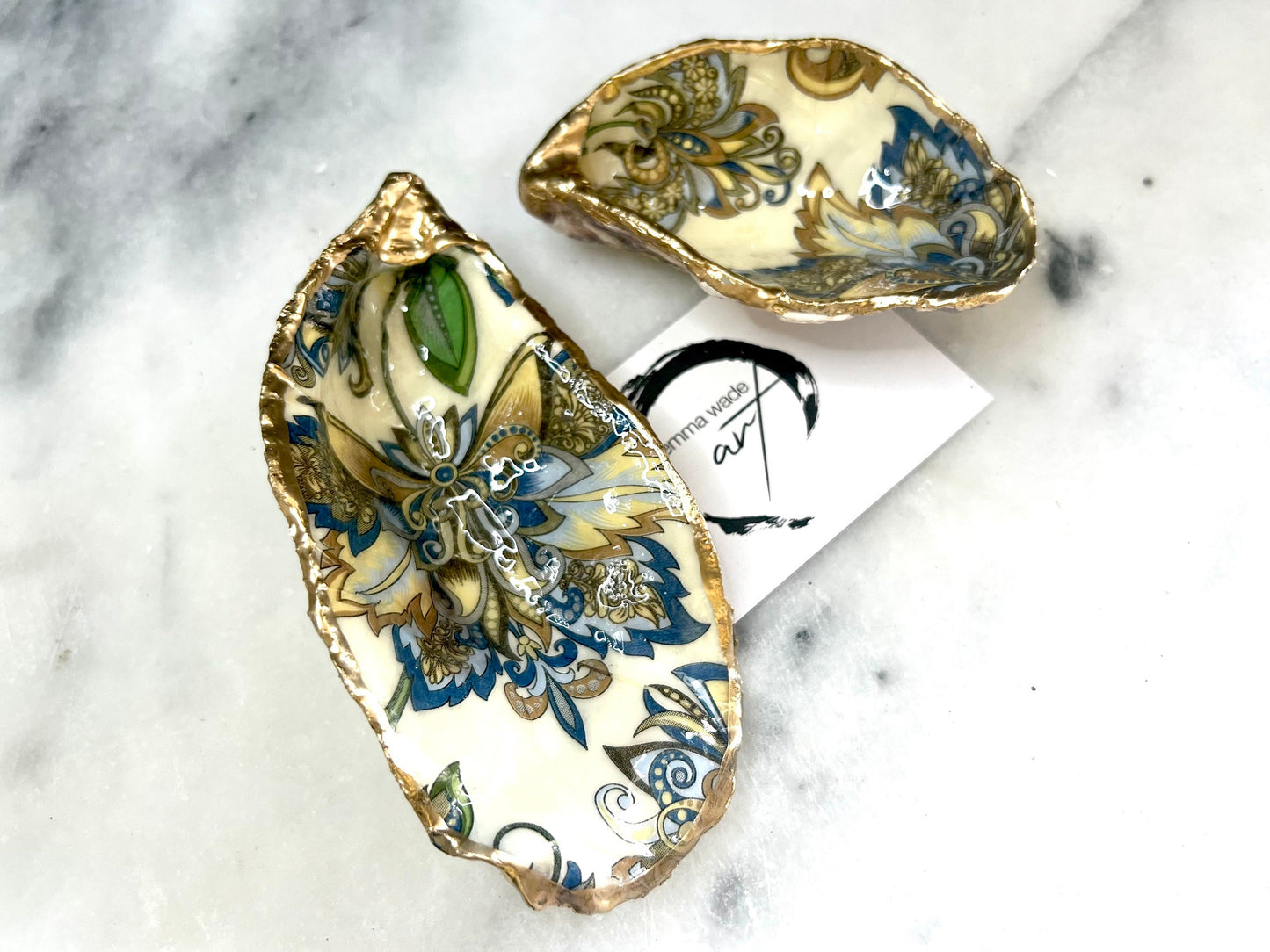 Handmade Dorset oyster shell trinket dishes decorated with ornate blue, green and gold baroque floral patterns inspired by antique European porcelain and vintage decorative art. Natural oyster shells are hand gilded with liquid gold leaf edging and sealed with glossy crystal clear resin. Luxury coastal jewelry holders and vanity dishes photographed on marble background with elegant decorative styling.
