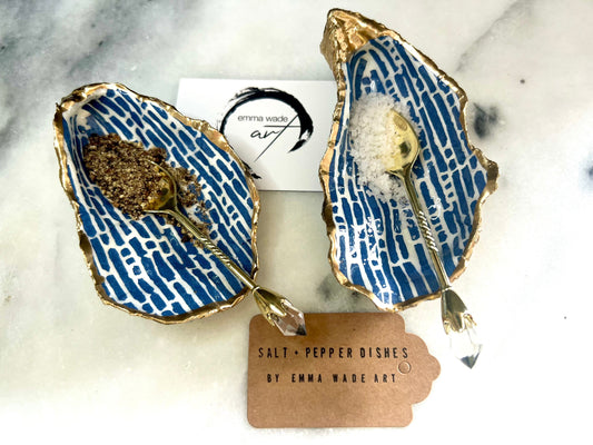 Pair of handmade blue and white lined oyster shell salt and pepper pinch pots with gold leaf trim, shown on a light background with small gold spoons. Crafted from natural Dorset oyster shells and sealed with glossy, food-safe resin, these coastal-inspired shell dishes are designed for table styling, weddings, or a unique hostess gift. Each shell is one of a kind and finished by hand.