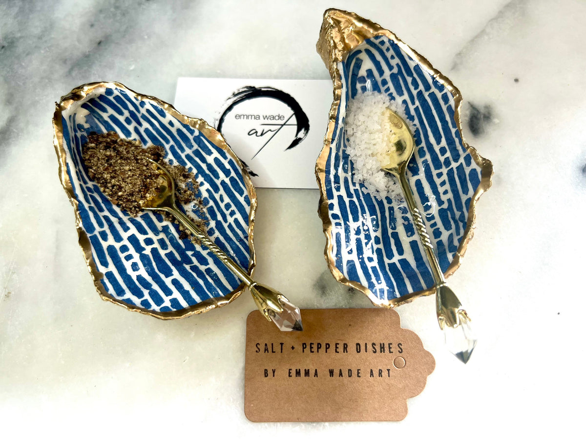 Pair of handmade blue and white lined oyster shell salt and pepper pinch pots with gold leaf trim, shown on a light background with small gold spoons. Crafted from natural Dorset oyster shells and sealed with glossy, food-safe resin, these coastal-inspired shell dishes are designed for table styling, weddings, or a unique hostess gift. Each shell is one of a kind and finished by hand.