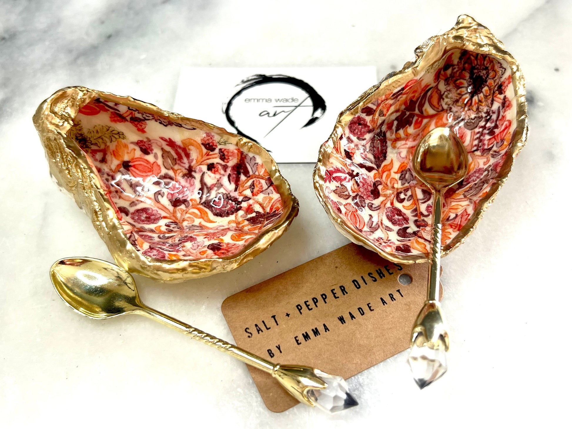 Handmade Dorset oyster shell salt and pepper pinch pots featuring a pink William Morris inspired floral design with hand-painted gold leaf edging. Decorative coastal tableware sealed in glossy food-safe resin and styled with gold crystal spoons on a marble background.