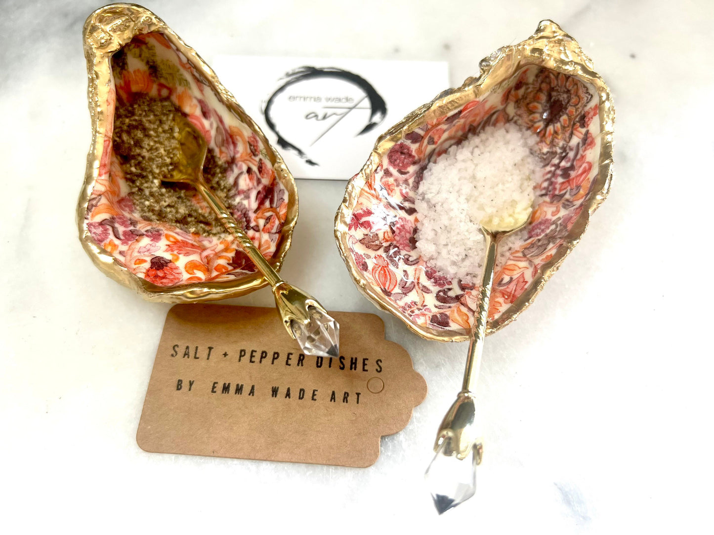Handmade Dorset oyster shell salt and pepper pinch pots featuring a pink William Morris inspired floral design with hand-painted gold leaf edging. Decorative coastal tableware sealed in glossy food-safe resin and styled with gold crystal spoons on a marble background.