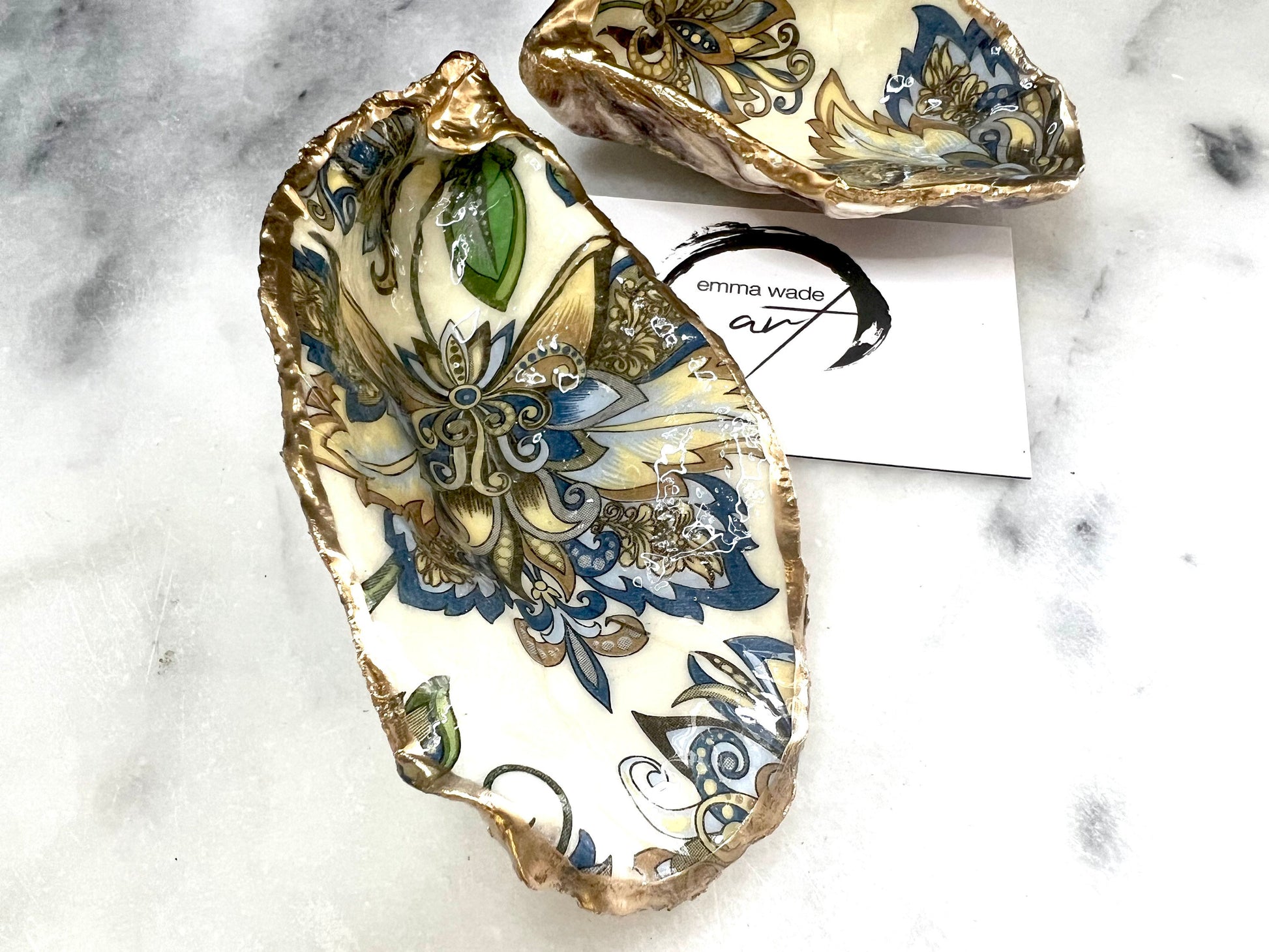 Handmade Dorset oyster shell trinket dishes decorated with ornate blue, green and gold baroque floral patterns inspired by antique European porcelain and vintage decorative art. Natural oyster shells are hand gilded with liquid gold leaf edging and sealed with glossy crystal clear resin. Luxury coastal jewelry holders and vanity dishes photographed on marble background with elegant decorative styling.