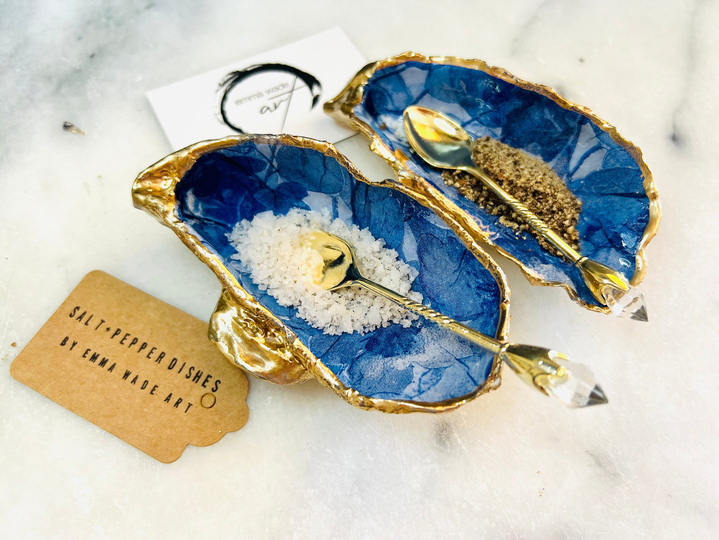 Pair of oyster shell salt and pepper dishes with deep blue watercolour flower design, finished with gold leaf trim and glossy seal. Handmade from Dorset oyster shells, each dish is unique with natural variations. A beautiful oyster shell salt gift set, also available with matching gold crystal spoons for a finishing touch. Perfect for weddings, anniversaries, dinner parties, or as a handmade gift for birthdays, Christmas, or housewarmings. Elegant coastal tableware and decor piece.