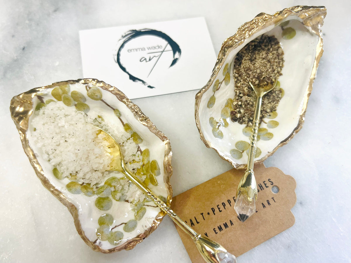 Pair of white and gold oyster shell salt and pepper pinch pots with delicate berry botanical design, hand gilded gold leaf edging and glossy resin finish. Handmade Dorset coastal shell dishes styled with gold crystal spoons on a marble background.