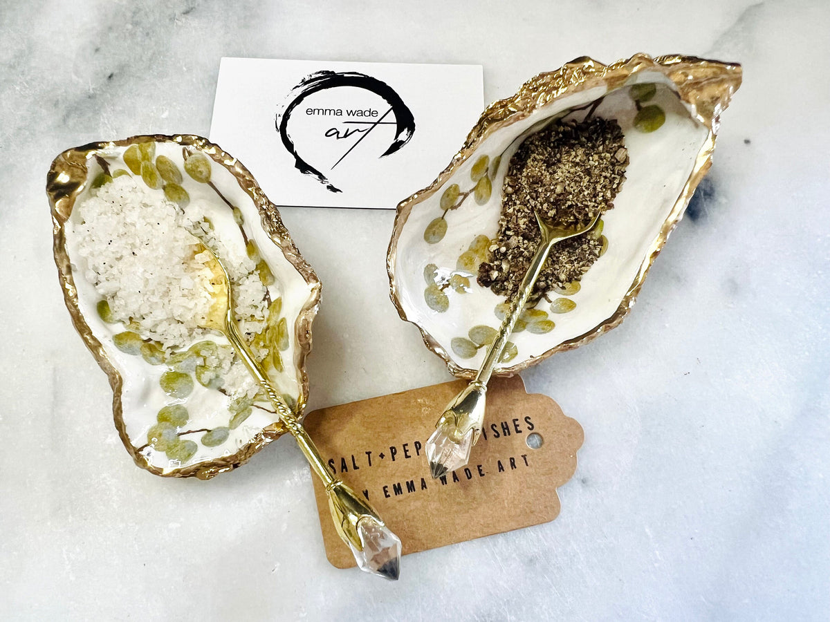 Pair of white and gold oyster shell salt and pepper pinch pots with delicate berry botanical design, hand gilded gold leaf edging and glossy resin finish. Handmade Dorset coastal shell dishes styled with gold crystal spoons on a marble background.