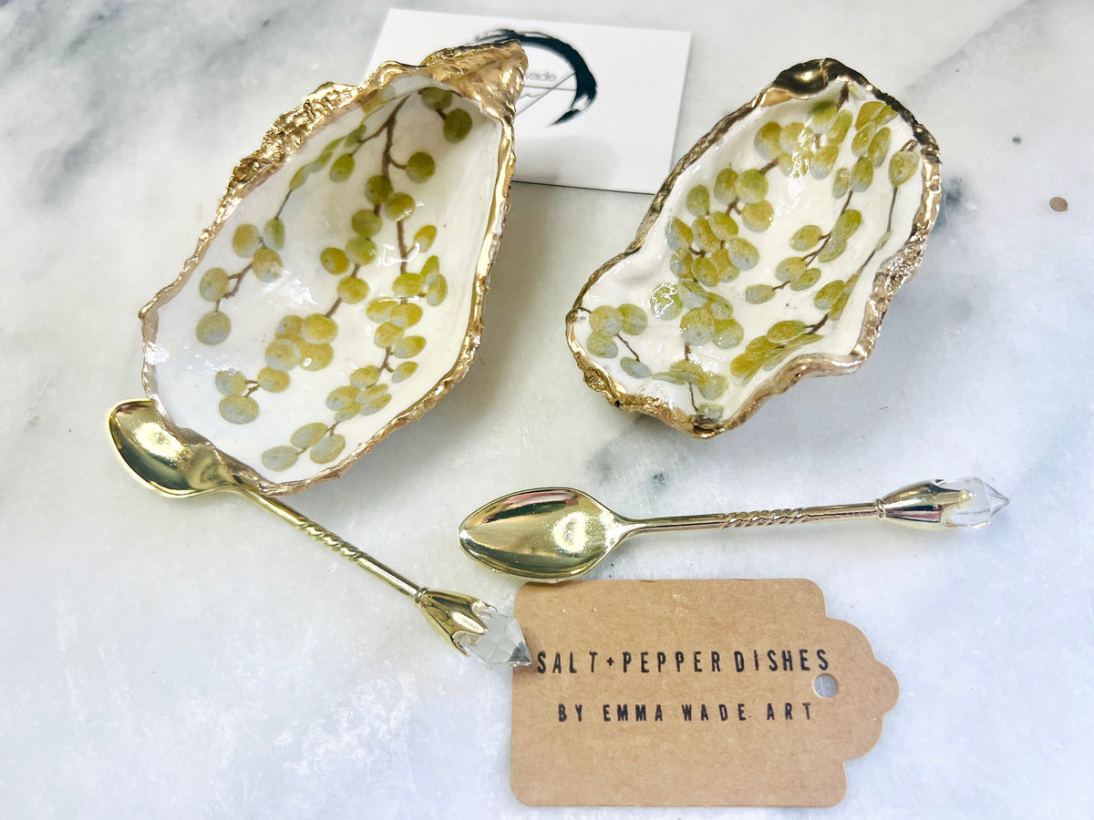 Pair of white and gold oyster shell salt and pepper pinch pots with delicate berry botanical design, hand gilded gold leaf edging and glossy resin finish. Handmade Dorset coastal shell dishes styled with gold crystal spoons on a marble background.