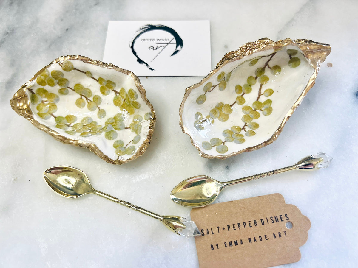 Pair of white and gold oyster shell salt and pepper pinch pots with delicate berry botanical design, hand gilded gold leaf edging and glossy resin finish. Handmade Dorset coastal shell dishes styled with gold crystal spoons on a marble background.