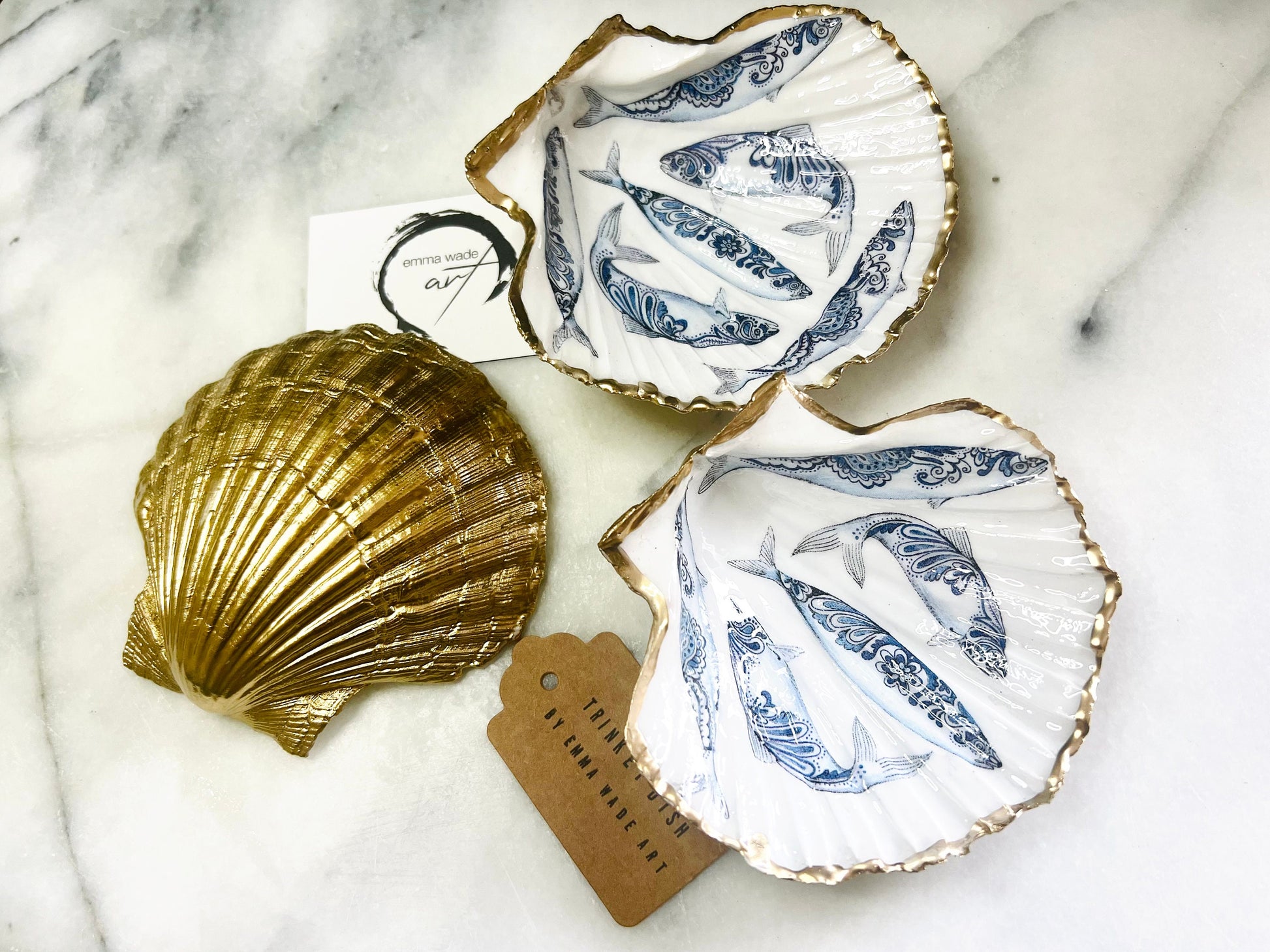 Handmade Dorset scallop shell trinket dishes featuring blue and white paisley fish illustrations with hand-painted gold leaf edging and glossy crystal clear resin. Coastal Scandinavian inspired jewellery dishes styled on a soft neutral background with elegant nautical fish artwork and luxurious gilded detailing.
