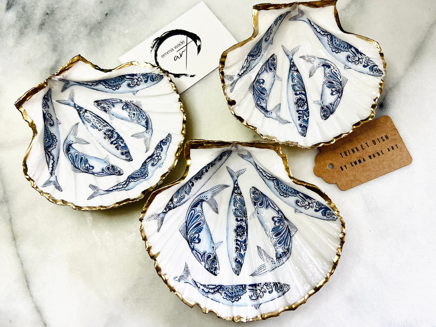 Handmade Dorset scallop shell trinket dishes featuring blue and white paisley fish illustrations with hand-painted gold leaf edging and glossy crystal clear resin. Coastal Scandinavian inspired jewellery dishes styled on a soft neutral background with elegant nautical fish artwork and luxurious gilded detailing.
