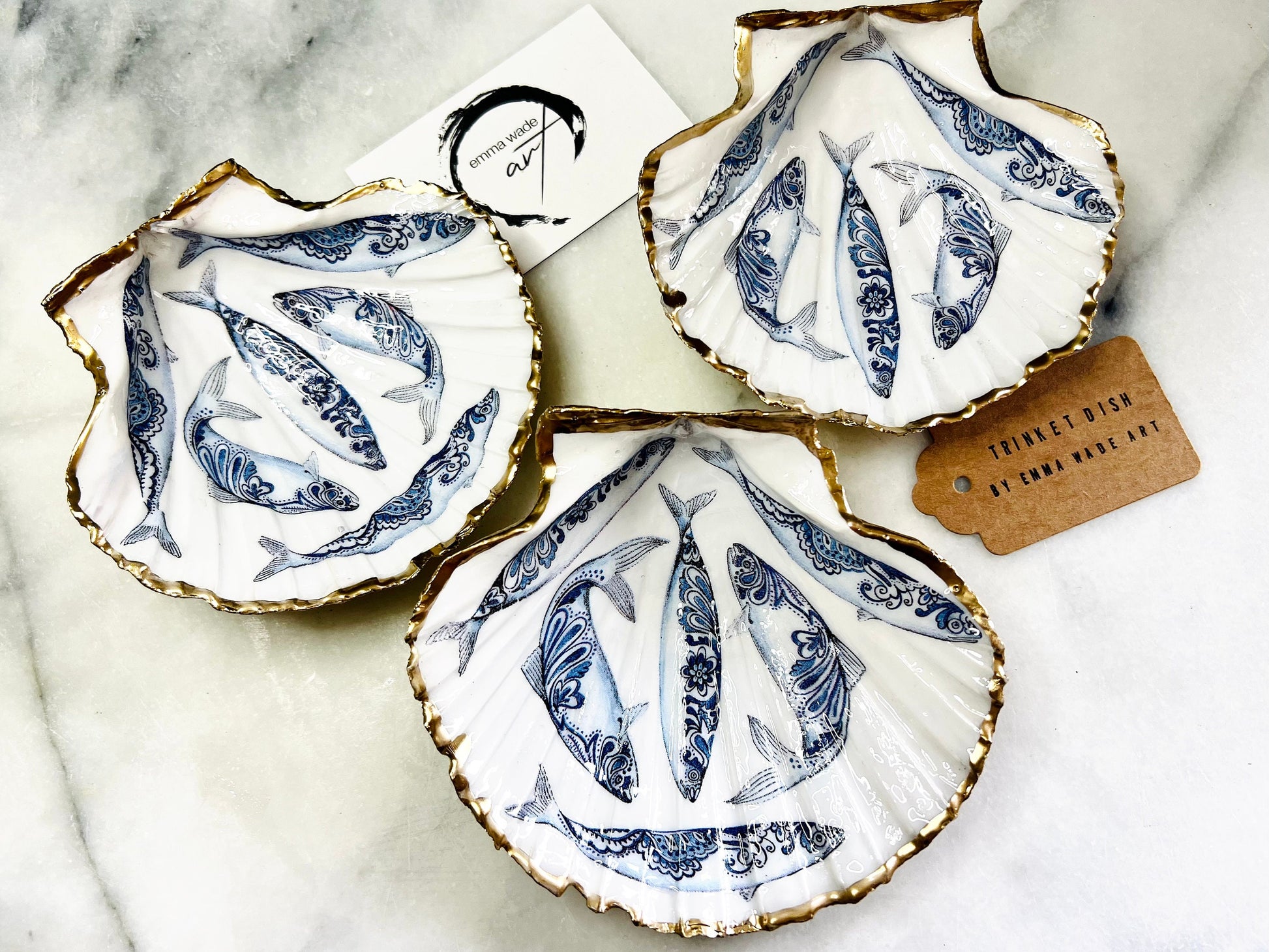 Handmade Dorset scallop shell trinket dishes featuring blue and white paisley fish illustrations with hand-painted gold leaf edging and glossy crystal clear resin. Coastal Scandinavian inspired jewellery dishes styled on a soft neutral background with elegant nautical fish artwork and luxurious gilded detailing.