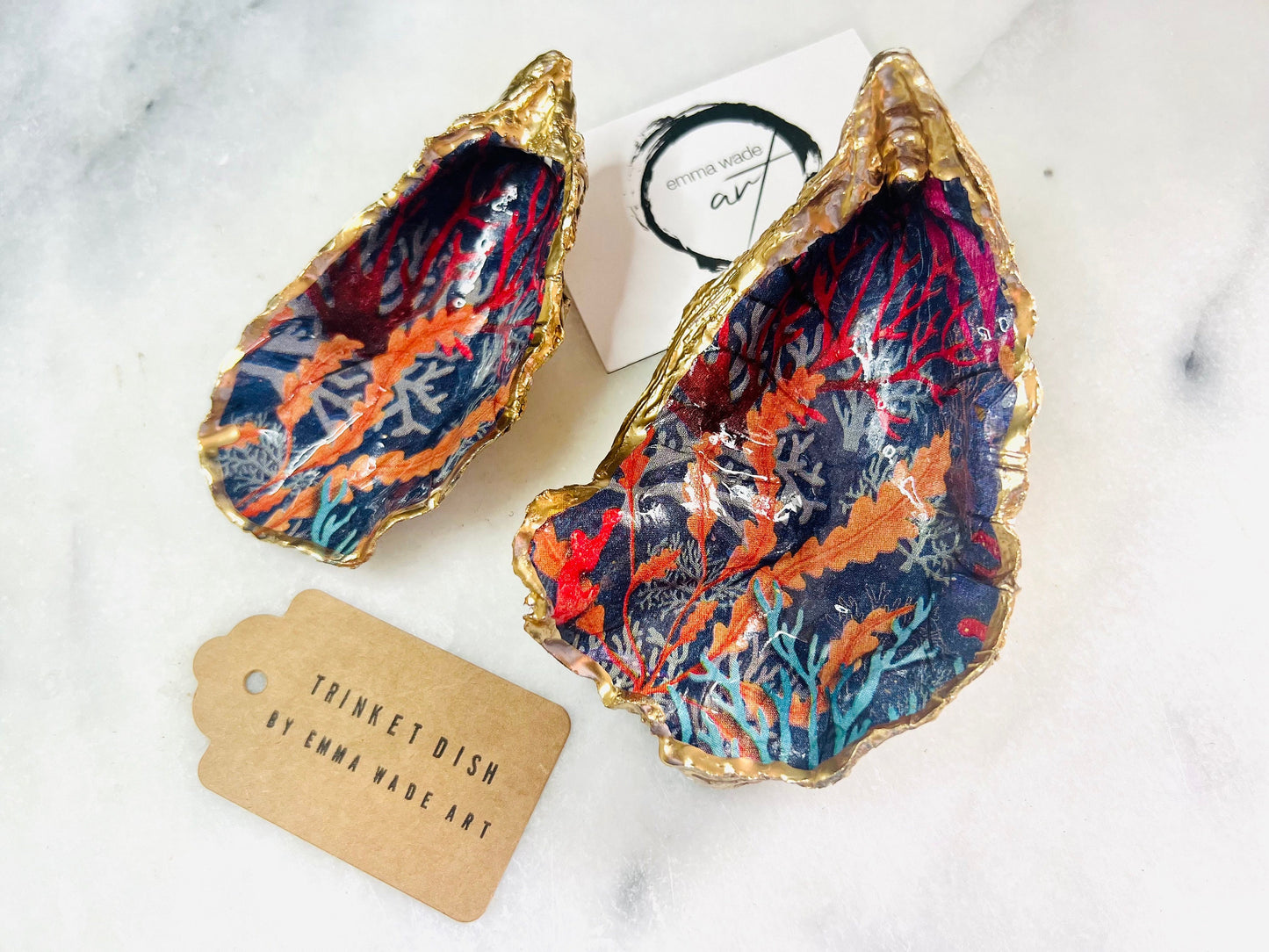 Handmade Dorset oyster shell trinket dishes featuring vibrant sea coral inspired artwork in coral red, navy and turquoise tones, finished with hand-painted gold leaf edging and glossy crystal clear resin. Coastal jewellery dishes styled on a white marble background with rich ocean-inspired botanical patterns and luxurious gilded edges.