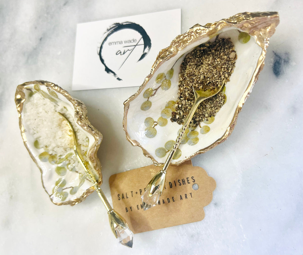 Pair of white and gold oyster shell salt and pepper pinch pots with delicate berry botanical design, hand gilded gold leaf edging and glossy resin finish. Handmade Dorset coastal shell dishes styled with gold crystal spoons on a marble background.