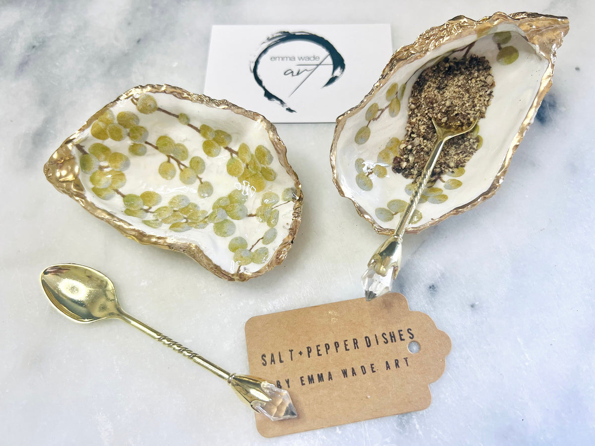 Pair of white and gold oyster shell salt and pepper pinch pots with delicate berry botanical design, hand gilded gold leaf edging and glossy resin finish. Handmade Dorset coastal shell dishes styled with gold crystal spoons on a marble background.