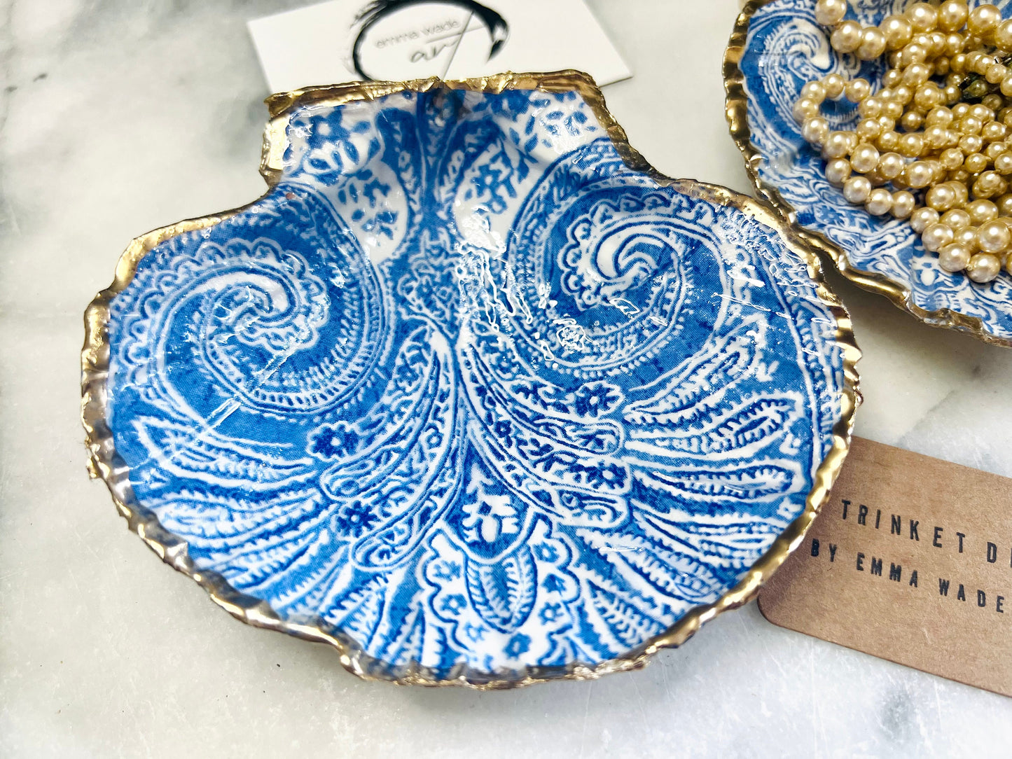 Blue and white paisley scallop shell ring catcher with hand applied gold leaf edging and glossy resin finish. Handmade Dorset shell jewellery tray styled with pearls on a marble background. Coastal vanity decor and luxury beach house accessory gift.