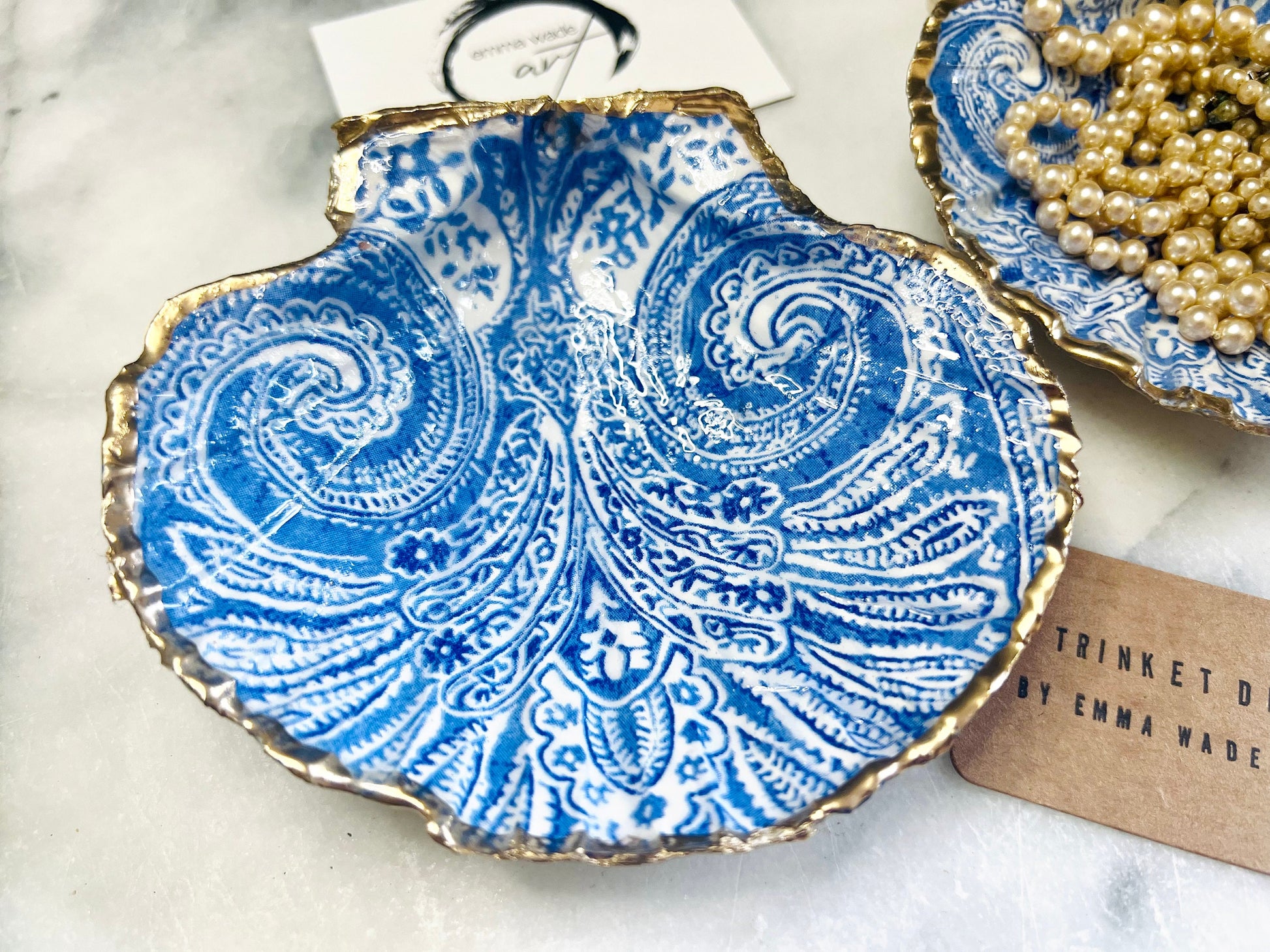 Blue and white paisley scallop shell ring catcher with hand applied gold leaf edging and glossy resin finish. Handmade Dorset shell jewellery tray styled with pearls on a marble background. Coastal vanity decor and luxury beach house accessory gift.