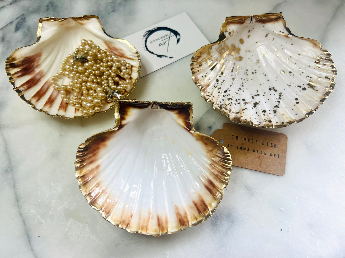 Handmade Dorset scallop shell trinket dishes featuring natural white shells with delicate liquid gold leaf speckling, hand-painted gold leaf edging and glossy crystal clear resin. Luxury coastal jewellery dishes styled on a white marble background with elegant neutral tones and artisan coastal home décor styling.