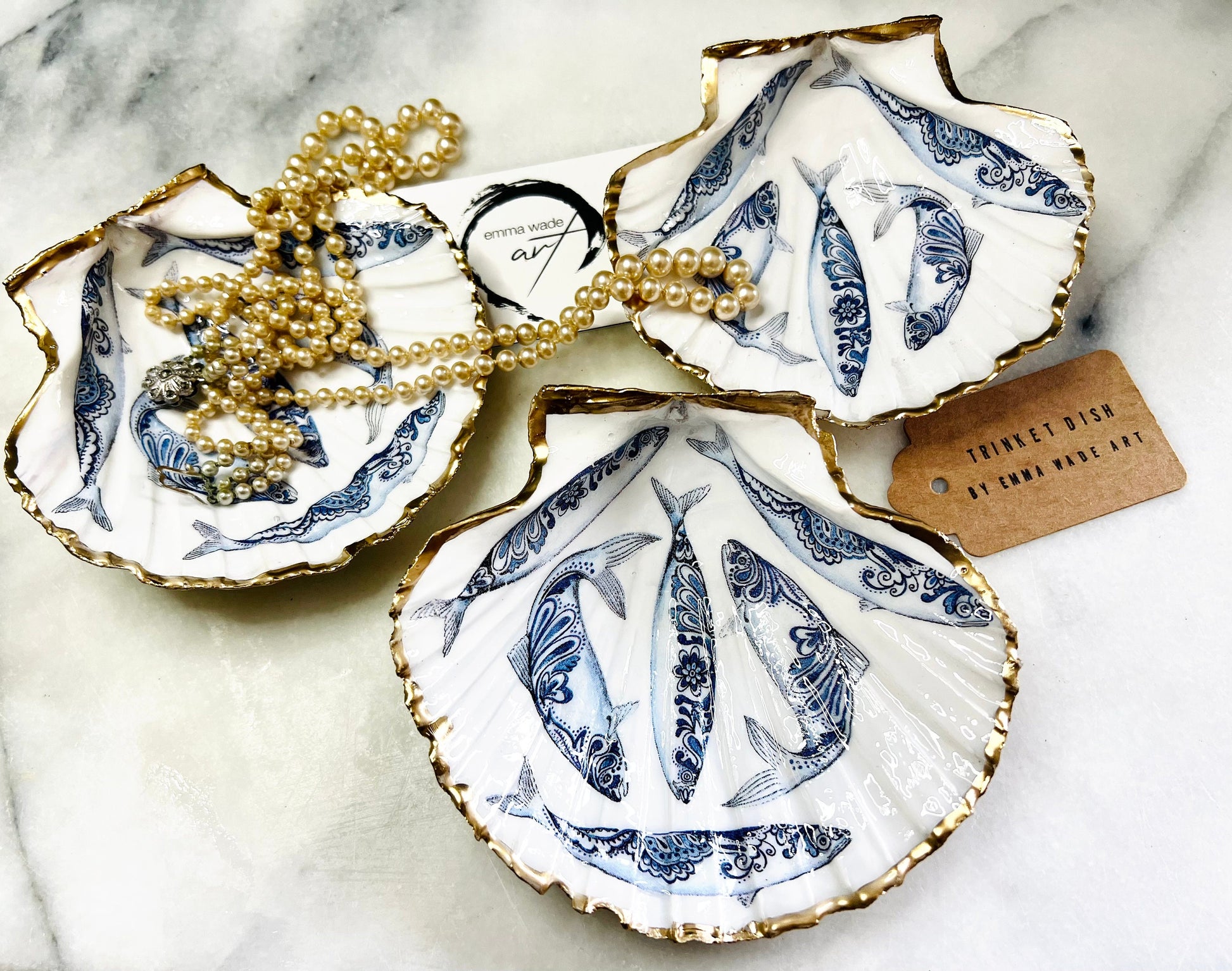Handmade Dorset scallop shell trinket dishes featuring blue and white paisley fish illustrations with hand-painted gold leaf edging and glossy crystal clear resin. Coastal Scandinavian inspired jewellery dishes styled on a soft neutral background with elegant nautical fish artwork and luxurious gilded detailing.