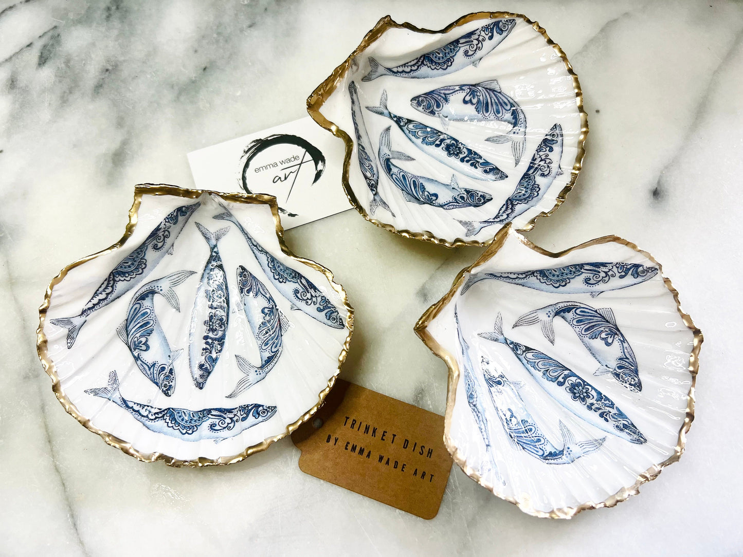 Handmade Dorset scallop shell trinket dishes featuring blue and white paisley fish illustrations with hand-painted gold leaf edging and glossy crystal clear resin. Coastal Scandinavian inspired jewellery dishes styled on a soft neutral background with elegant nautical fish artwork and luxurious gilded detailing.