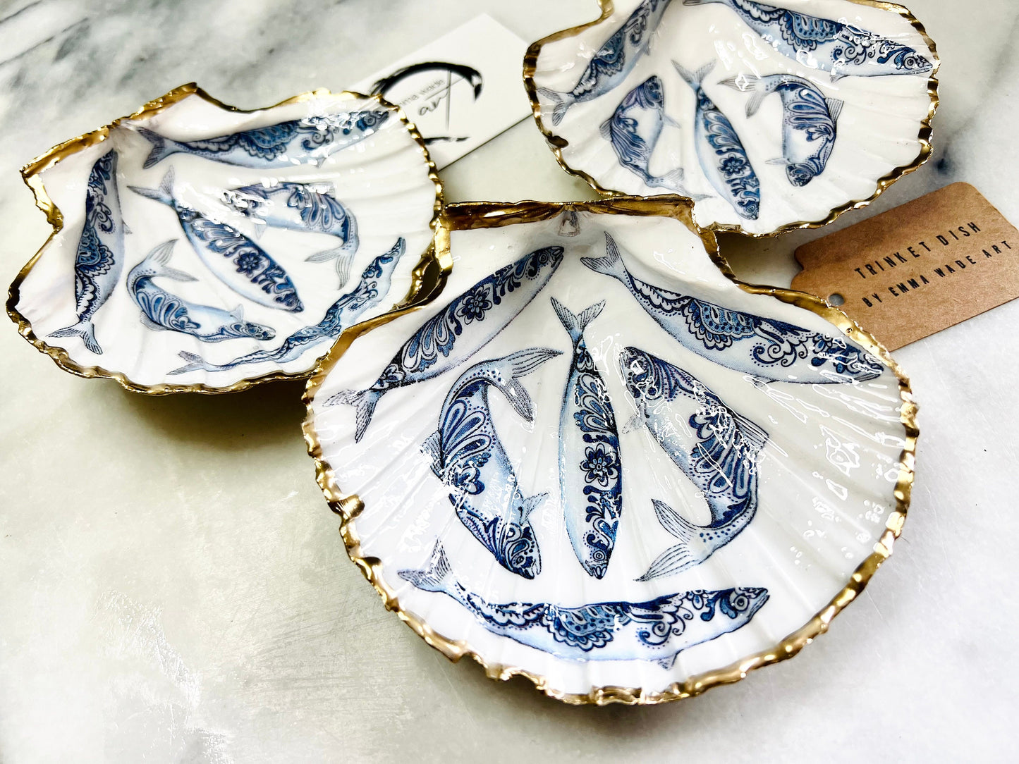 Handmade Dorset scallop shell trinket dishes featuring blue and white paisley fish illustrations with hand-painted gold leaf edging and glossy crystal clear resin. Coastal Scandinavian inspired jewellery dishes styled on a soft neutral background with elegant nautical fish artwork and luxurious gilded detailing.
