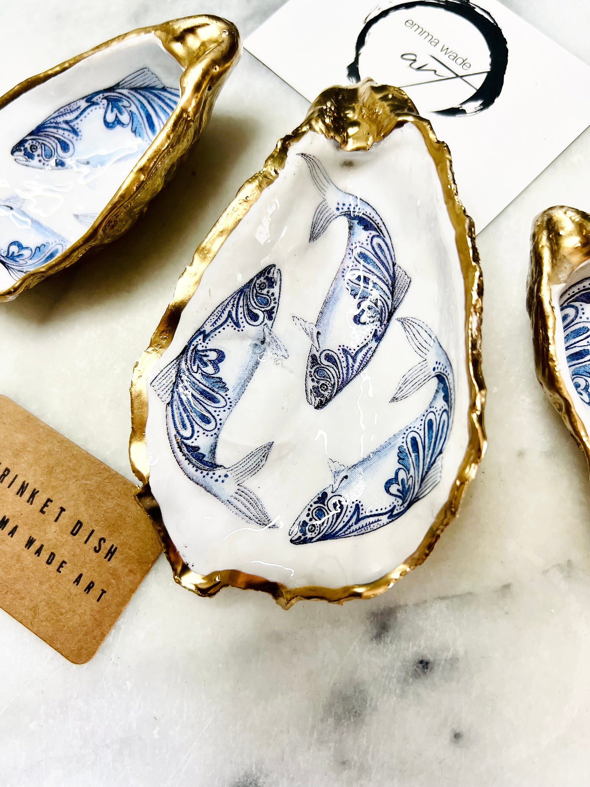 Handmade Dorset oyster shell trinket dish with blue and white paisley fish design, gold leaf gilded edges and glossy resin finish. Coastal jewellery holder and ring dish, photographed on a light surface showing the natural shell shape and pearly interior. Unique sustainably sourced shell, sold individually as a decorative coastal home décor accent or gift.