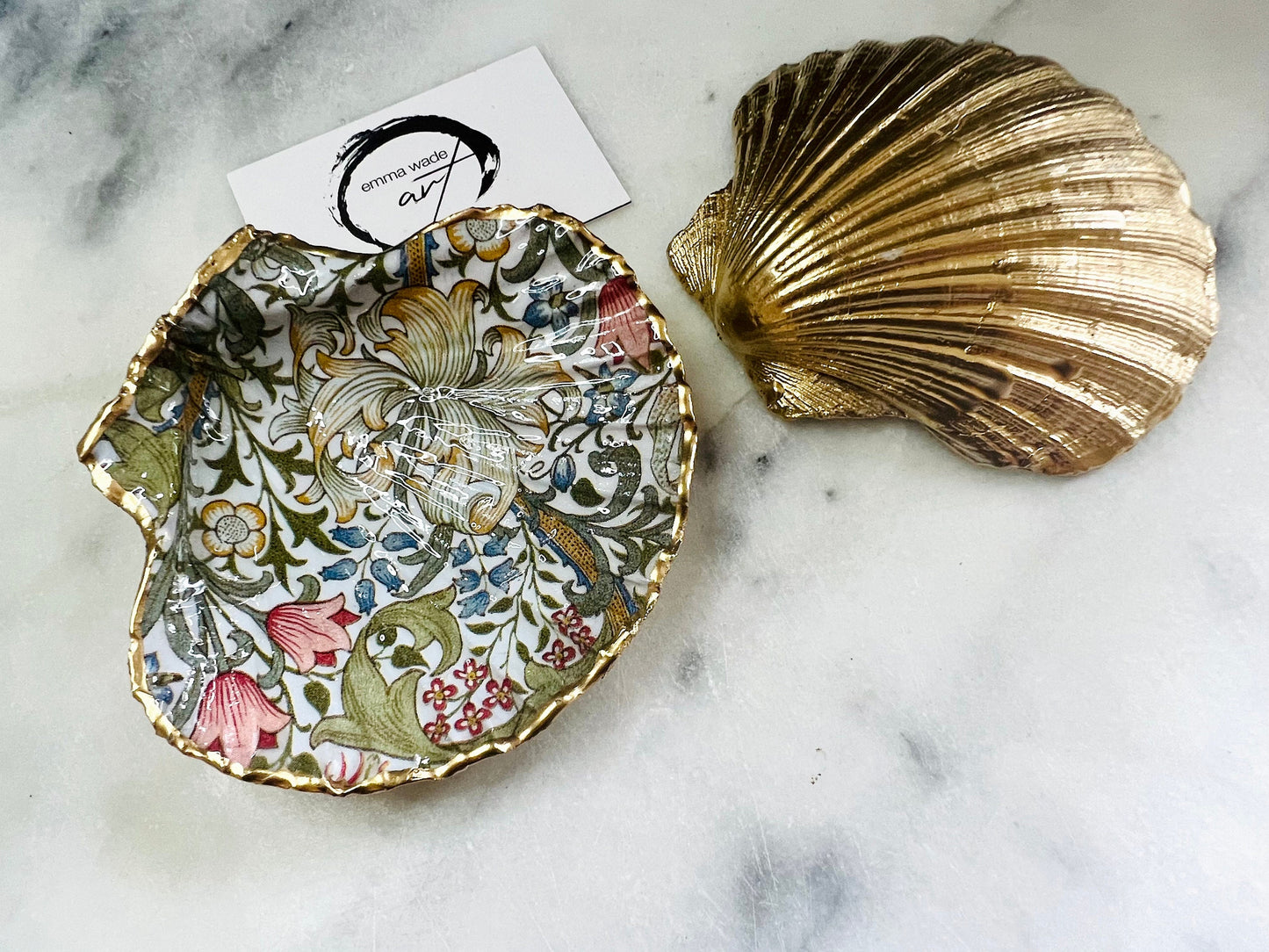 Handcrafted Dorset scallop shell trinket dish featuring William Morris Golden Lily floral design. Natural shell edged with hand applied gold leaf and sealed with glossy resin. Heritage floral jewellery holder displayed with rings on marble surface. Elegant coastal home decor gift.