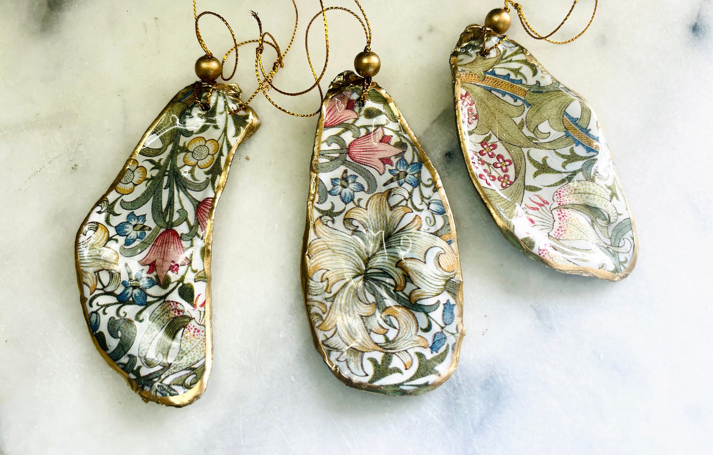 Handmade Dorset oyster shell hanging ornaments featuring William Morris inspired Golden Lily and Granville floral designs in soft green, blush pink and gold tones. Natural oyster shells finished with liquid gold leaf edging and sealed in glossy resin. Decorative hanging ornaments with gold cord and bead detail, suitable for luxury Christmas décor, coastal interiors, cottagecore styling or elegant year-round home decoration.