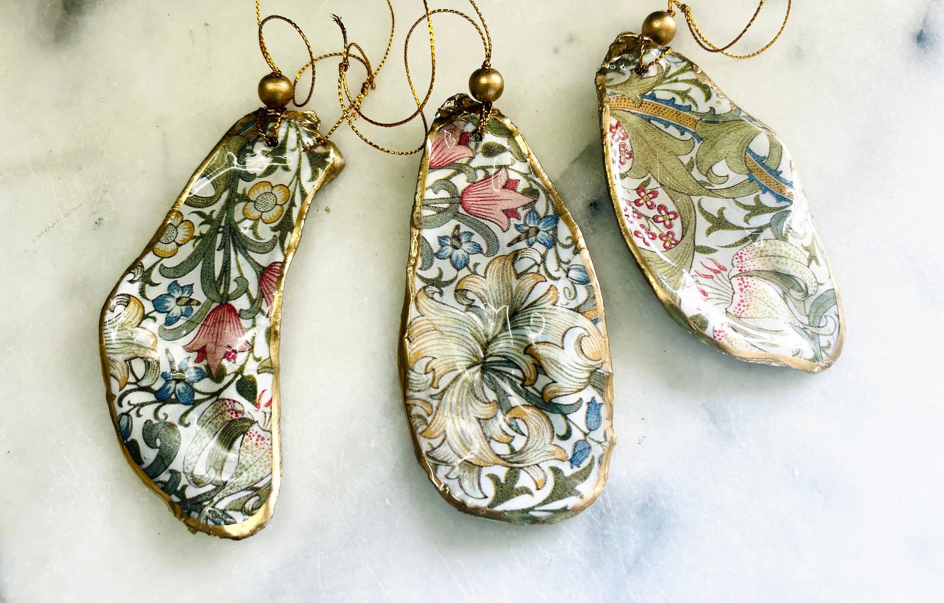 Handmade Dorset oyster shell hanging ornaments featuring William Morris inspired Golden Lily and Granville floral designs in soft green, blush pink and gold tones. Natural oyster shells finished with liquid gold leaf edging and sealed in glossy resin. Decorative hanging ornaments with gold cord and bead detail, suitable for luxury Christmas décor, coastal interiors, cottagecore styling or elegant year-round home decoration.