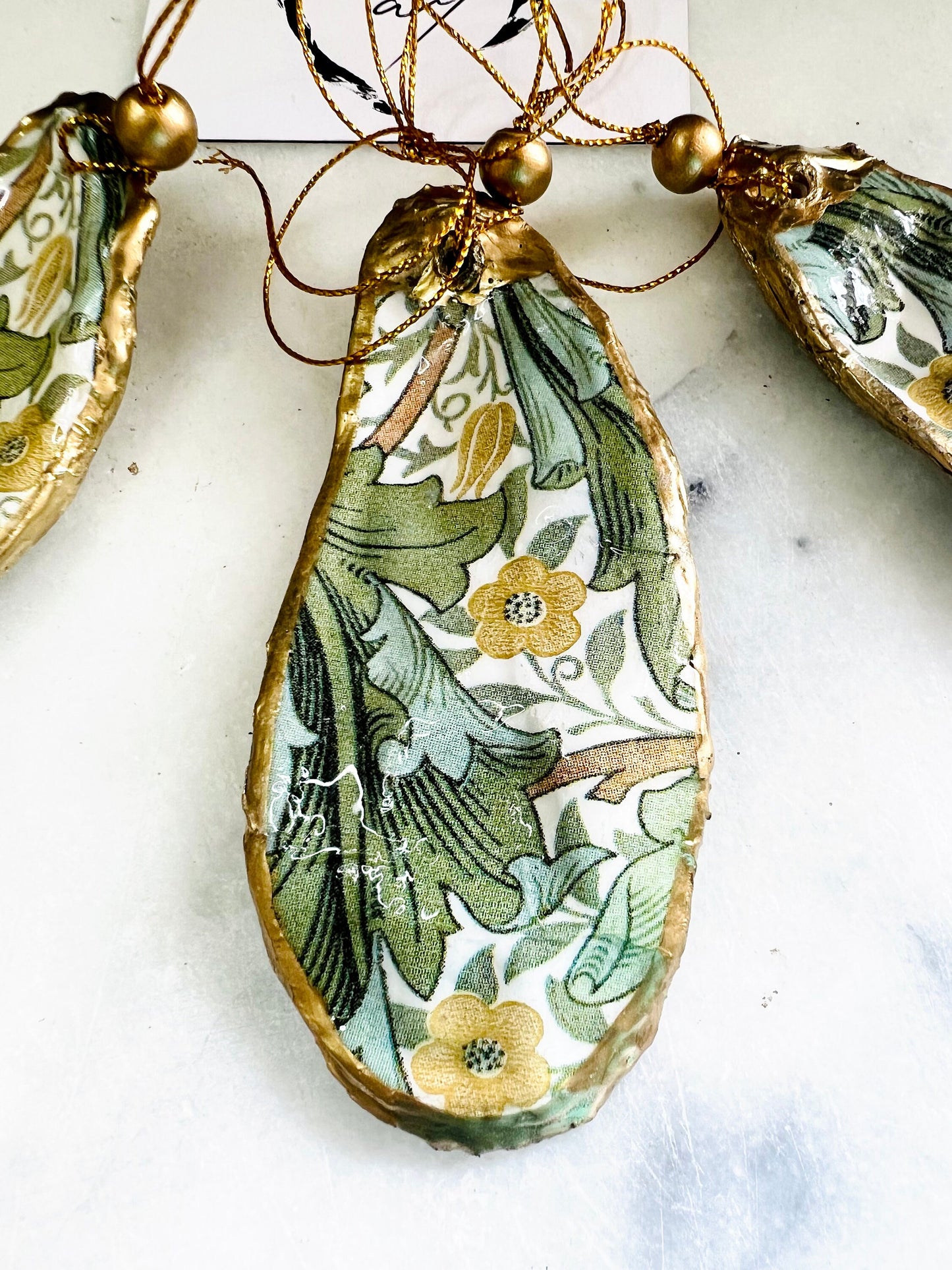 Handmade Dorset oyster shell ornaments decorated with William Morris Gold Granville and J.H. Dearle Golden Lily designs. Each shell features a natural pearl interior, gold leaf edging, and glossy resin seal. Pre-drilled with gold string and bead for hanging. Options available: individual ornament, matching set of 3, or mixed set of 6. Unique handmade shell decorations perfect for Christmas tree ornaments, wedding favours, bridesmaid gifts, or as William Morris style coastal-inspired home decor.