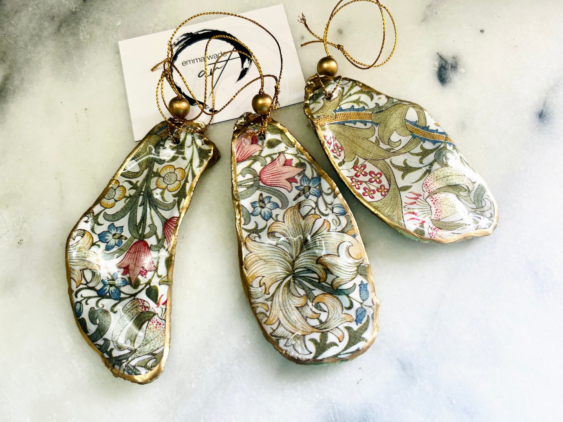 Handmade Dorset oyster shell hanging ornaments featuring William Morris inspired Golden Lily and Granville floral designs in soft green, blush pink and gold tones. Natural oyster shells finished with liquid gold leaf edging and sealed in glossy resin. Decorative hanging ornaments with gold cord and bead detail, suitable for luxury Christmas décor, coastal interiors, cottagecore styling or elegant year-round home decoration.