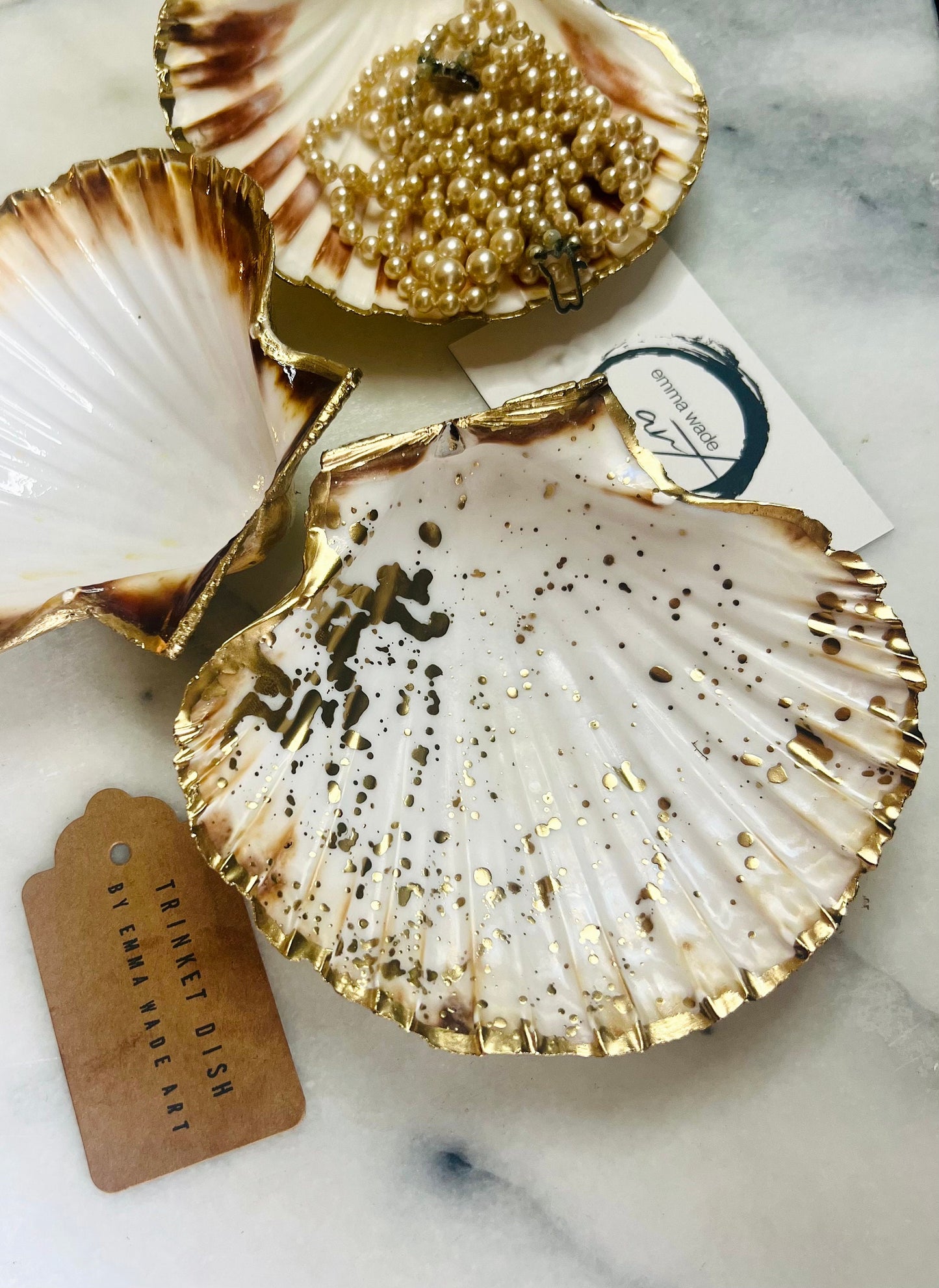 Handmade Dorset scallop shell trinket dishes featuring natural white shells with delicate liquid gold leaf speckling, hand-painted gold leaf edging and glossy crystal clear resin. Luxury coastal jewellery dishes styled on a white marble background with elegant neutral tones and artisan coastal home décor styling.