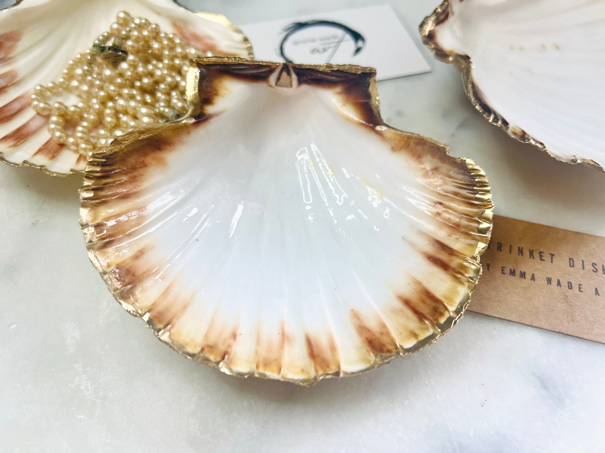 Handmade Dorset scallop shell trinket dishes featuring natural white shells with delicate liquid gold leaf speckling, hand-painted gold leaf edging and glossy crystal clear resin. Luxury coastal jewellery dishes styled on a white marble background with elegant neutral tones and artisan coastal home décor styling.