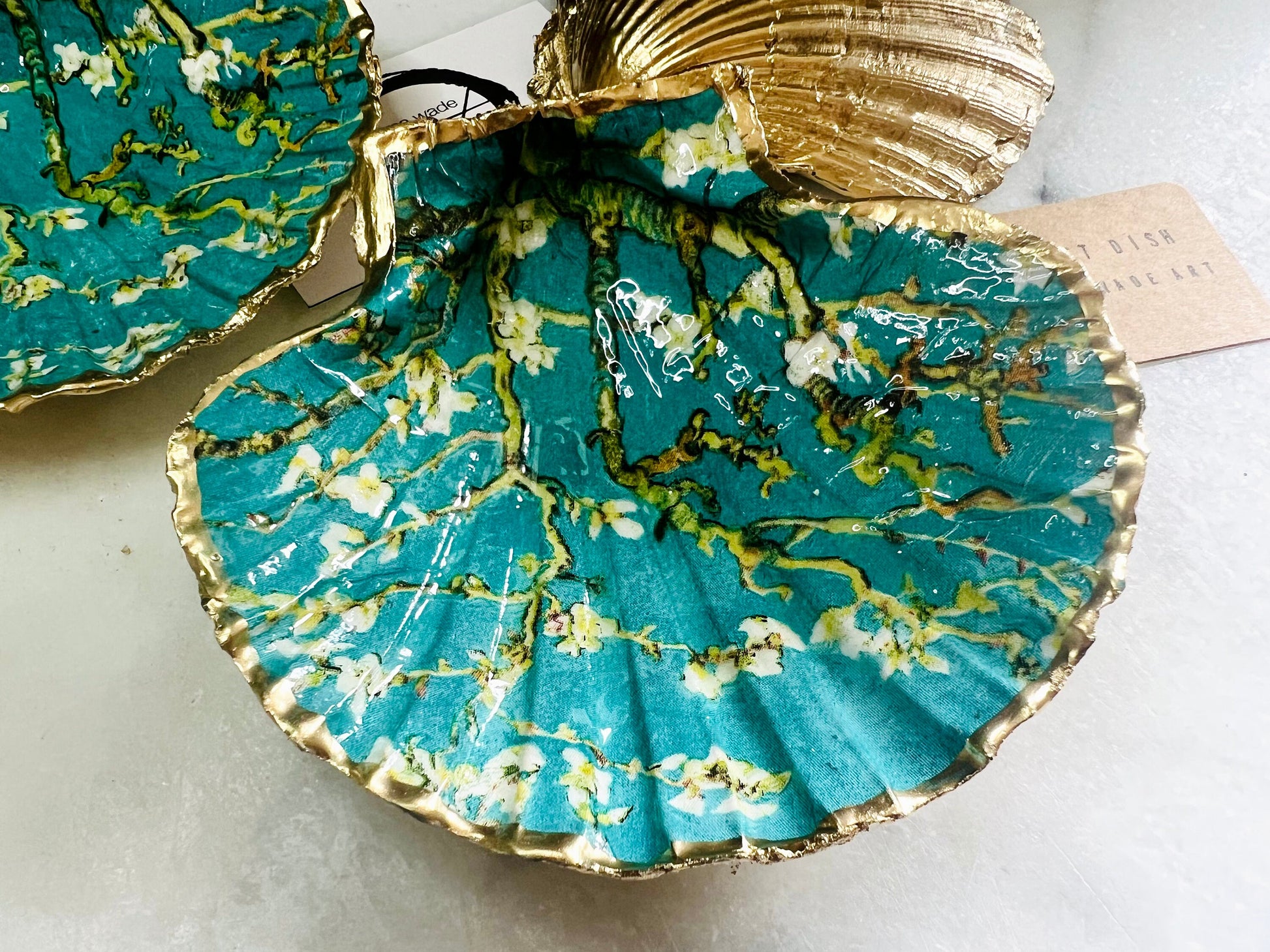 Handcrafted Dorset scallop shell trinket dish inspired by Vincent van Gogh’s Almond Blossom painting. Turquoise blue and green background with branching blossom design and raised gold detailing, finished with liquid gold leaf edging and sealed in high-gloss resin. Decorative shell jewellery holder styled on marble surface, suitable for rings, earrings, trinkets or as a tea light holder. Coastal art-inspired home decor piece.