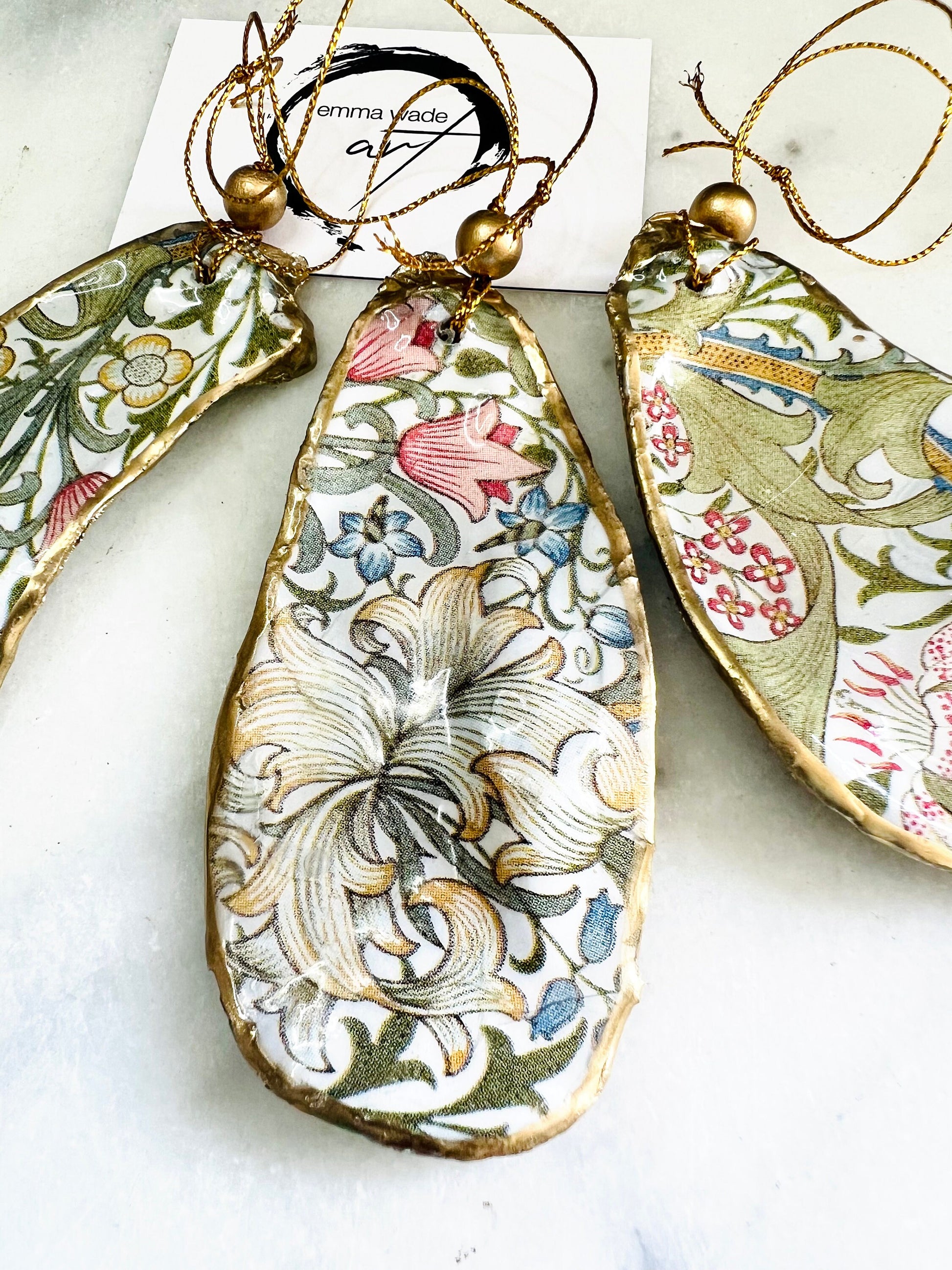 Handmade Dorset oyster shell hanging ornaments featuring William Morris inspired Golden Lily and Granville floral designs in soft green, blush pink and gold tones. Natural oyster shells finished with liquid gold leaf edging and sealed in glossy resin. Decorative hanging ornaments with gold cord and bead detail, suitable for luxury Christmas décor, coastal interiors, cottagecore styling or elegant year-round home decoration.