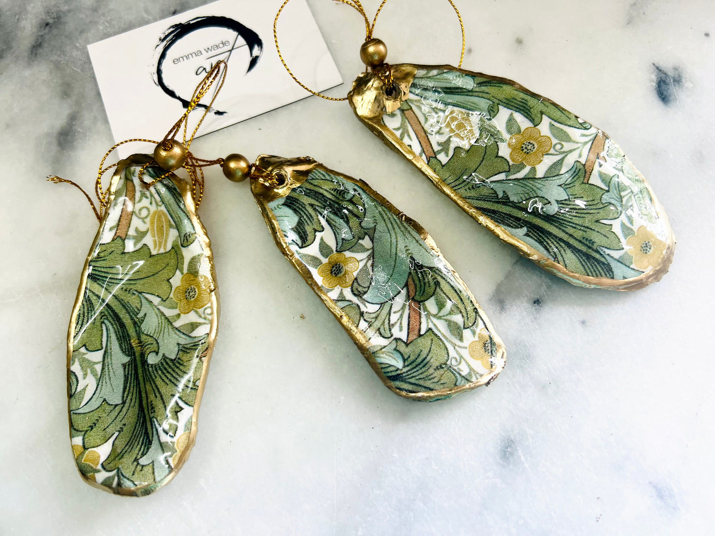 Handmade Dorset oyster shell hanging ornaments featuring William Morris inspired Golden Lily and Granville floral designs in soft green, blush pink and gold tones. Natural oyster shells finished with liquid gold leaf edging and sealed in glossy resin. Decorative hanging ornaments with gold cord and bead detail, suitable for luxury Christmas décor, coastal interiors, cottagecore styling or elegant year-round home decoration.