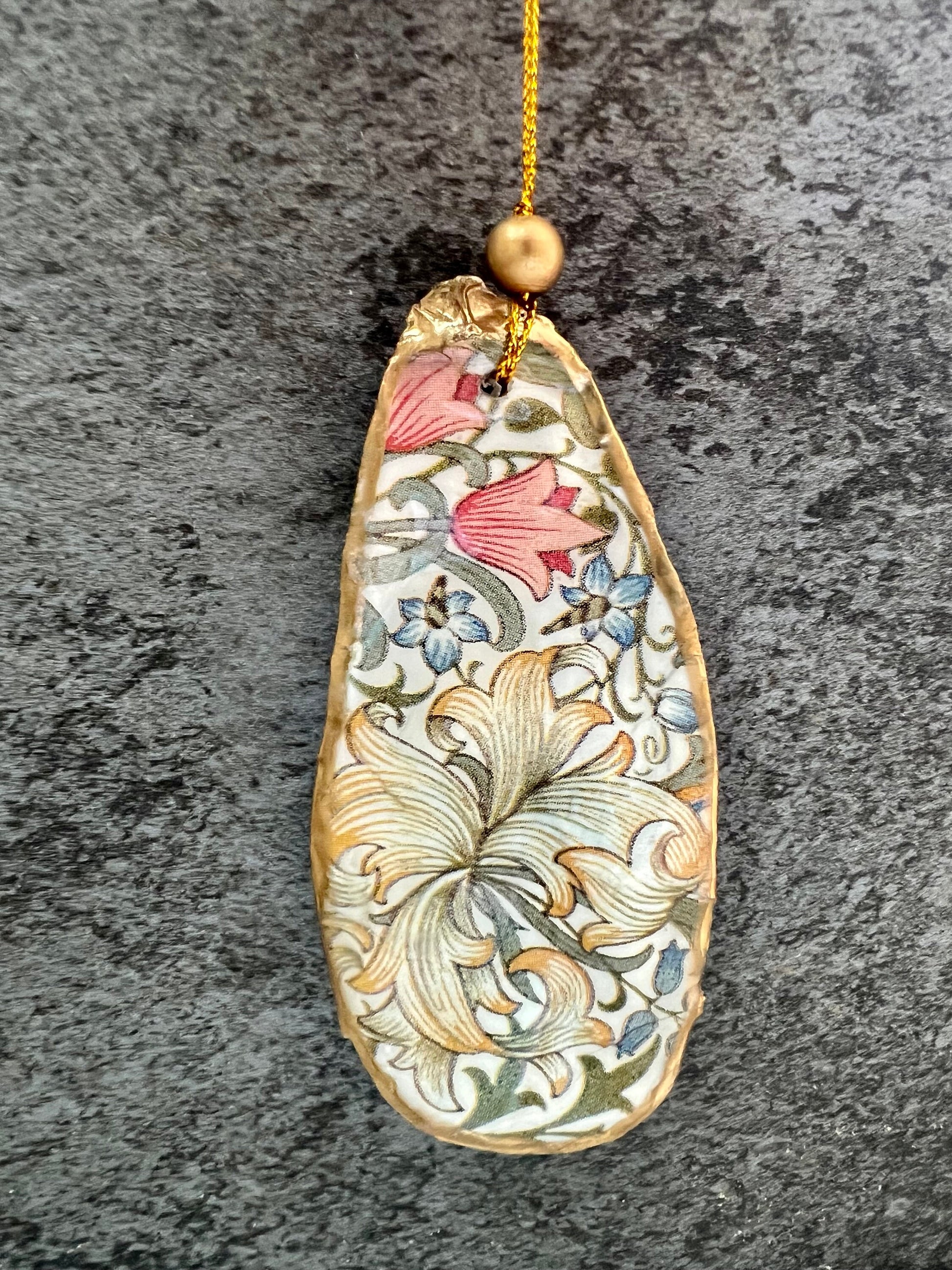 Handmade Dorset oyster shell hanging ornaments featuring William Morris inspired Golden Lily and Granville floral designs in soft green, blush pink and gold tones. Natural oyster shells finished with liquid gold leaf edging and sealed in glossy resin. Decorative hanging ornaments with gold cord and bead detail, suitable for luxury Christmas décor, coastal interiors, cottagecore styling or elegant year-round home decoration.